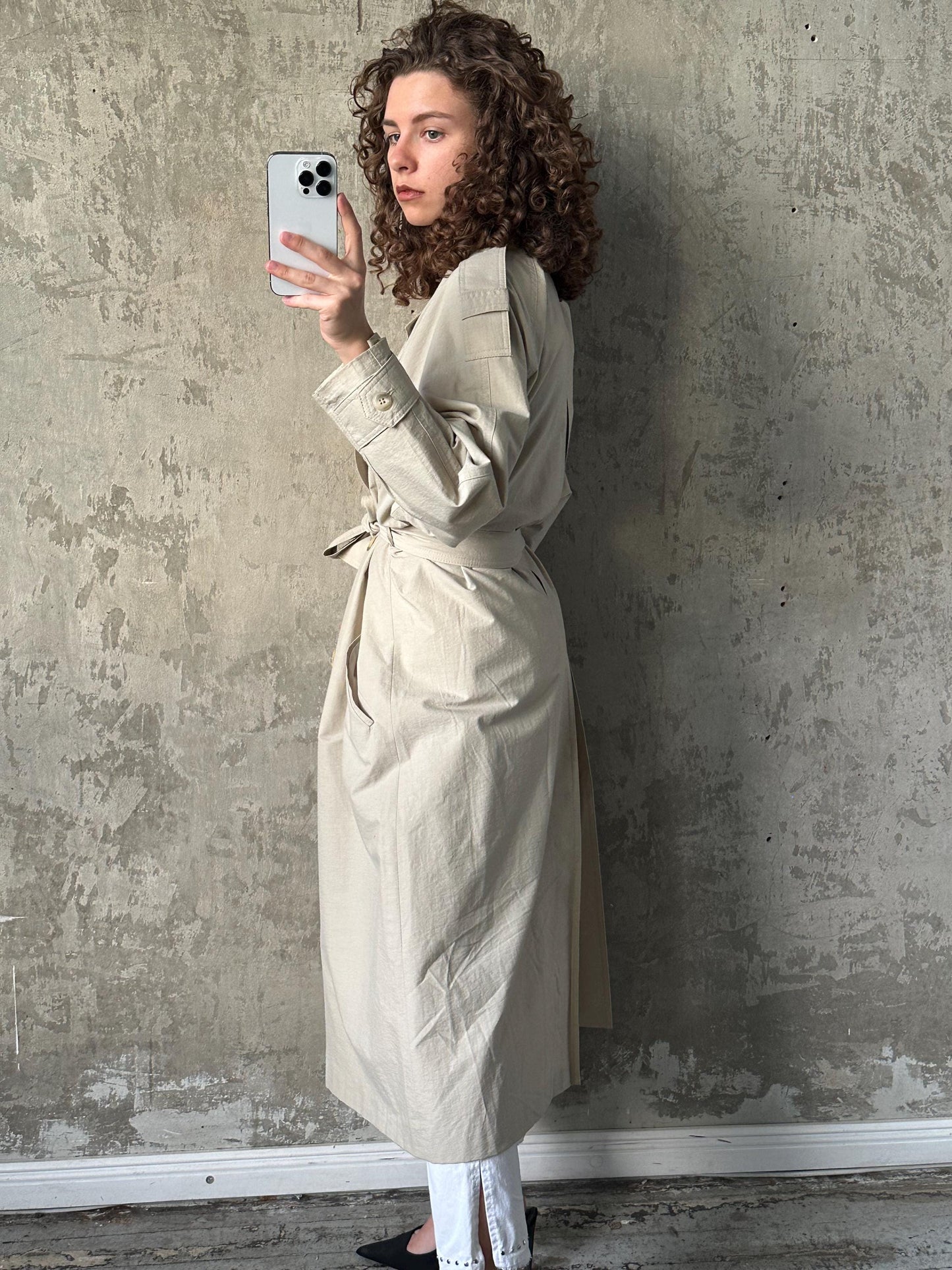 A woman with curly hair is taking a selfie with a smartphone, wearing a beige trench coat.