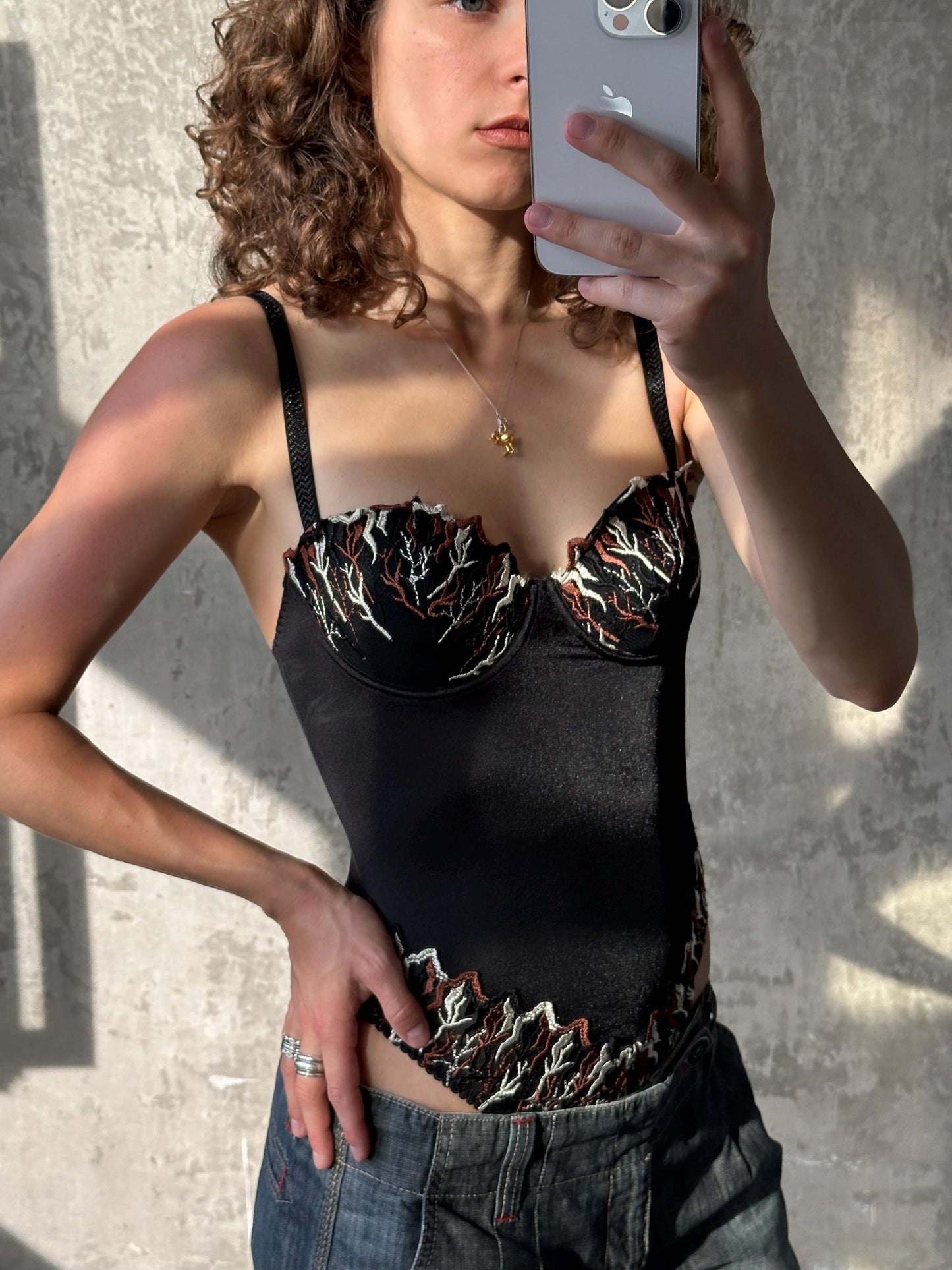 A woman with curly hair is taking a selfie with her iPhone, wearing a black corset with a floral pattern and jeans.
