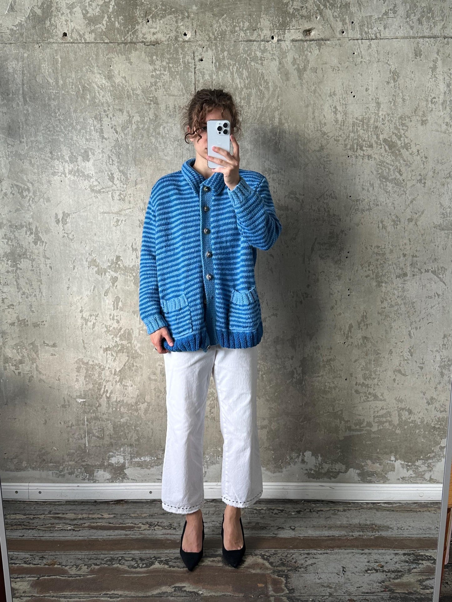 A person is taking a selfie with a blue striped cardigan and white pants, standing in front of a concrete wall.