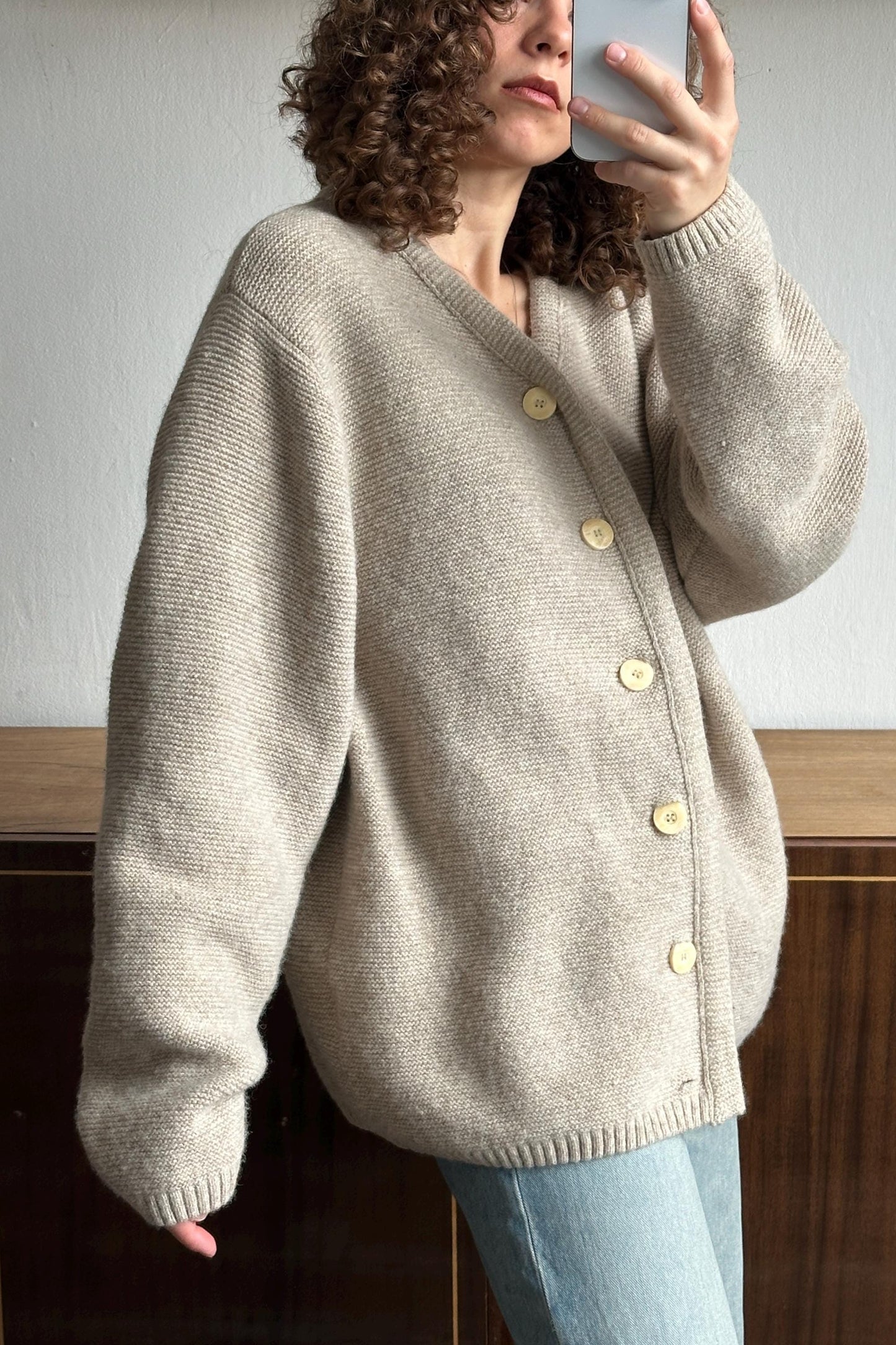 A woman with curly hair is taking a selfie with a smartphone, wearing a beige cardigan with buttons.