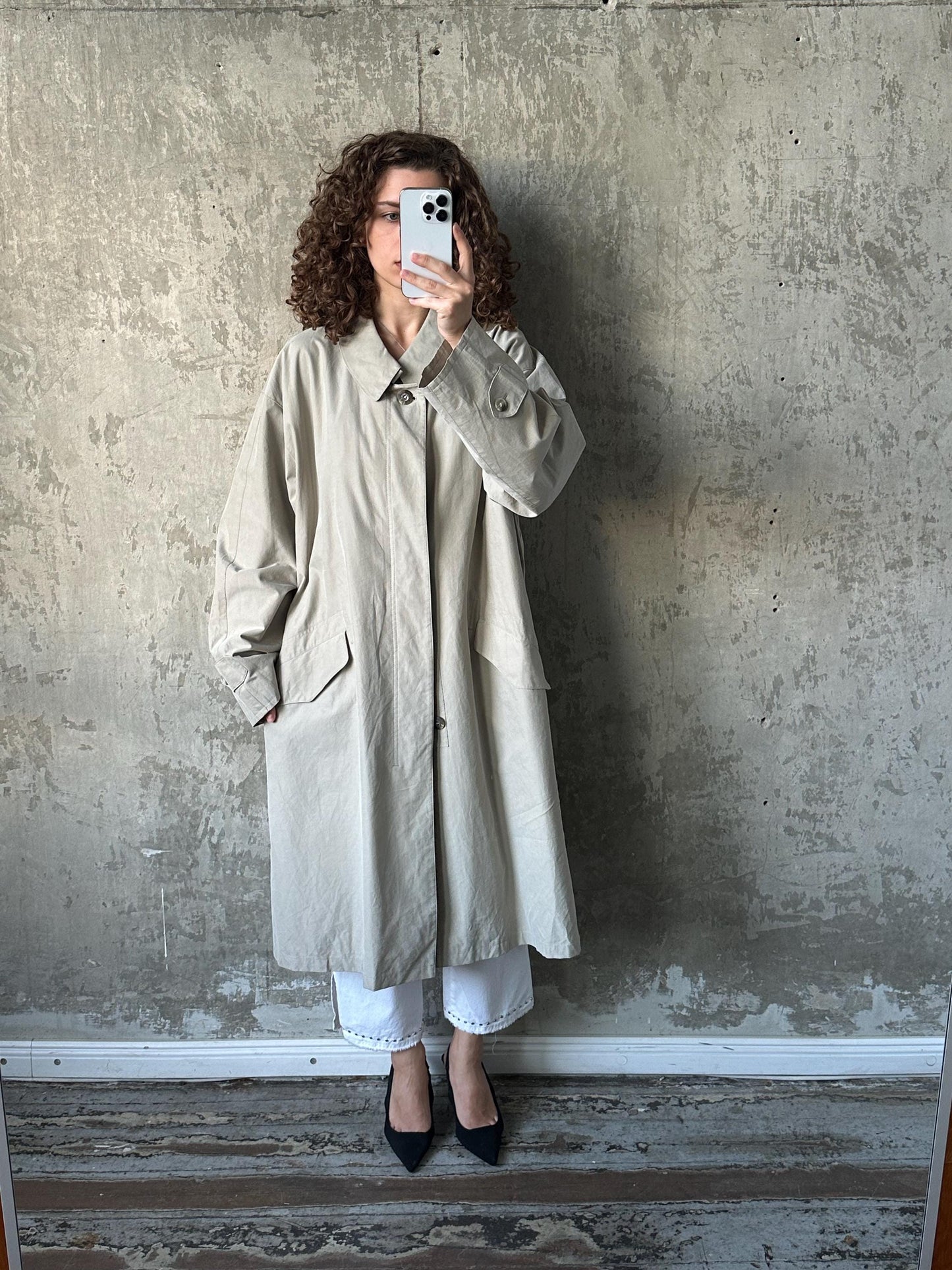 A woman in a beige trench coat is taking a selfie with her phone against a concrete wall.