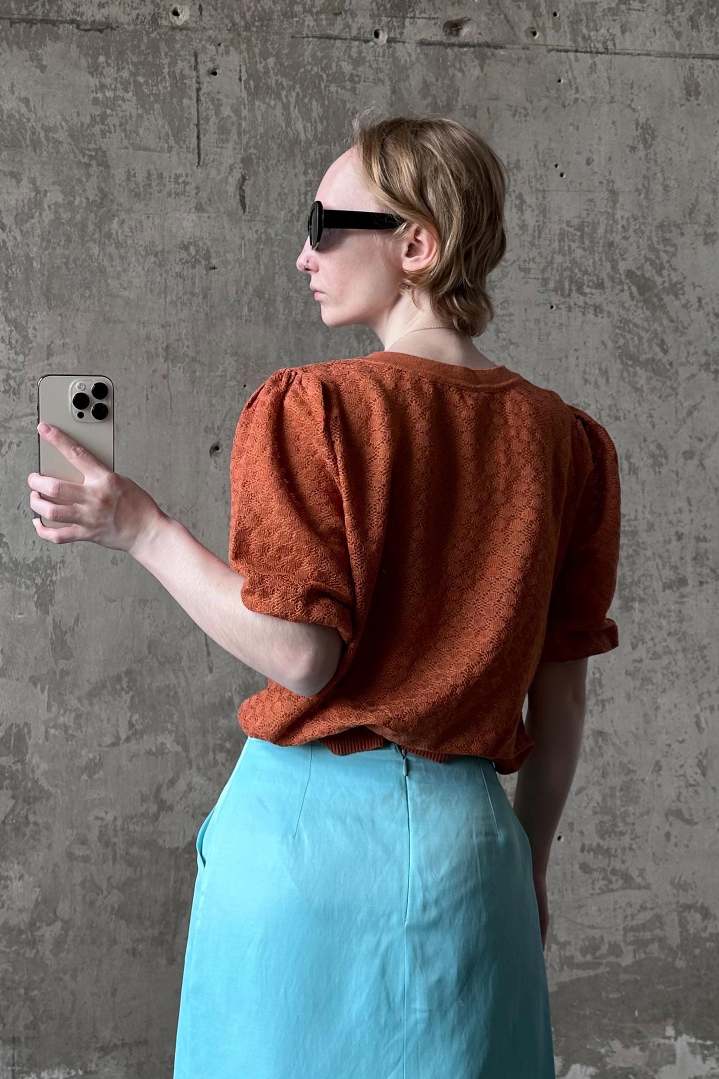 A woman in a red top and blue skirt is taking a selfie with a smartphone, holding the device up to her face.