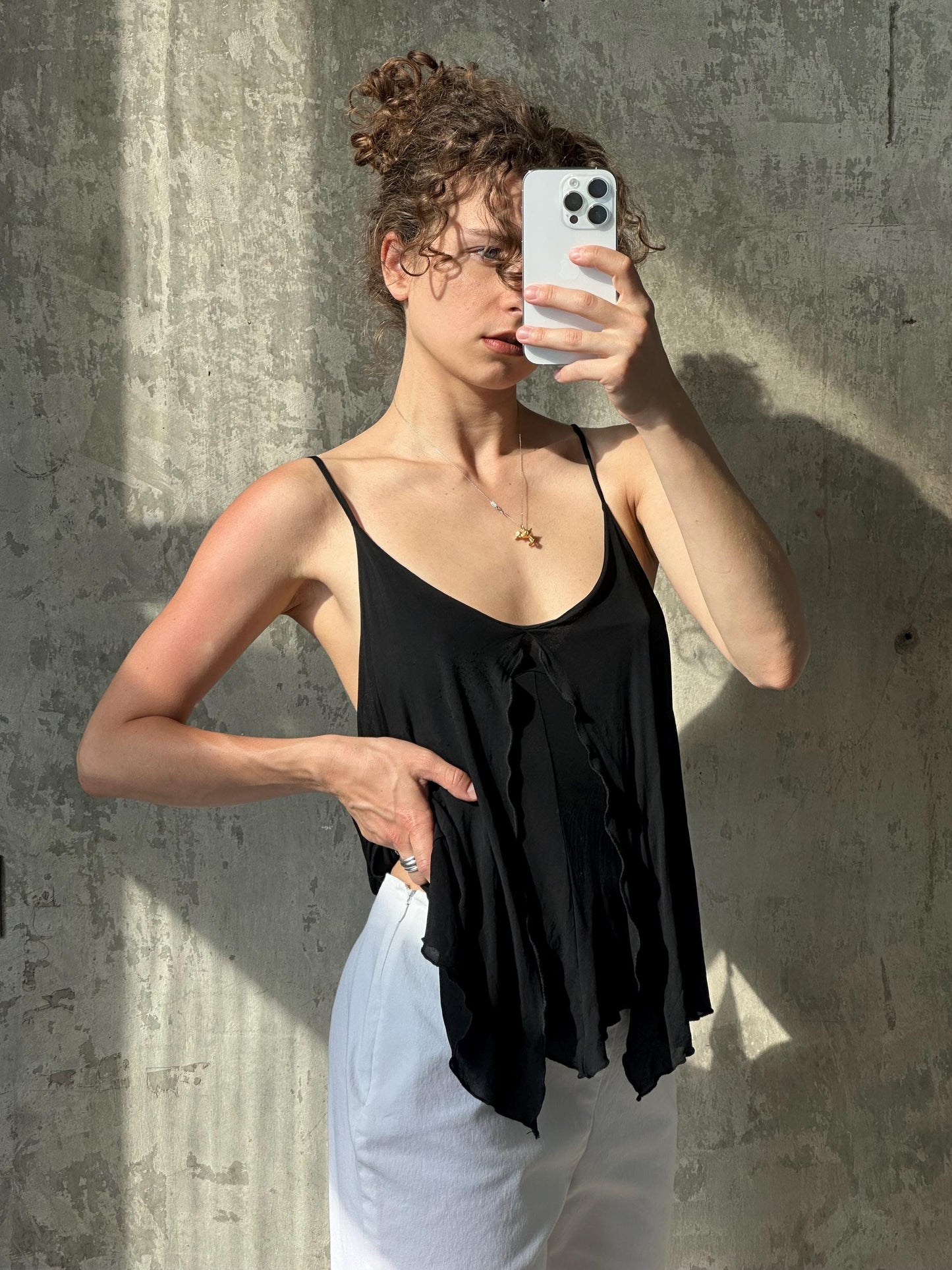 A woman with curly hair is taking a selfie with a white iPhone, holding the phone up to her face against a concrete wall.