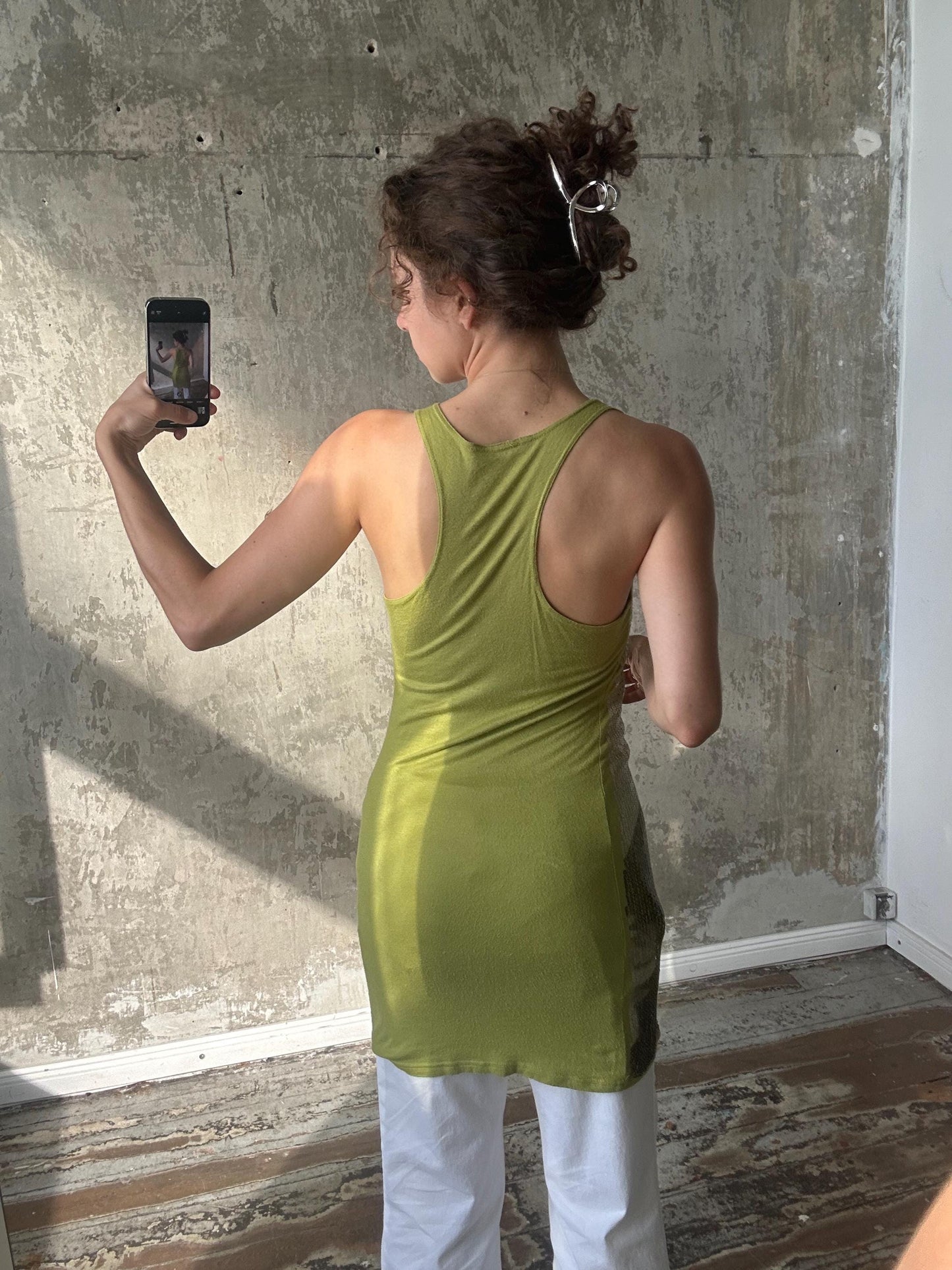 A woman in a green tank top is taking a selfie with a smartphone, standing in front of a concrete wall.