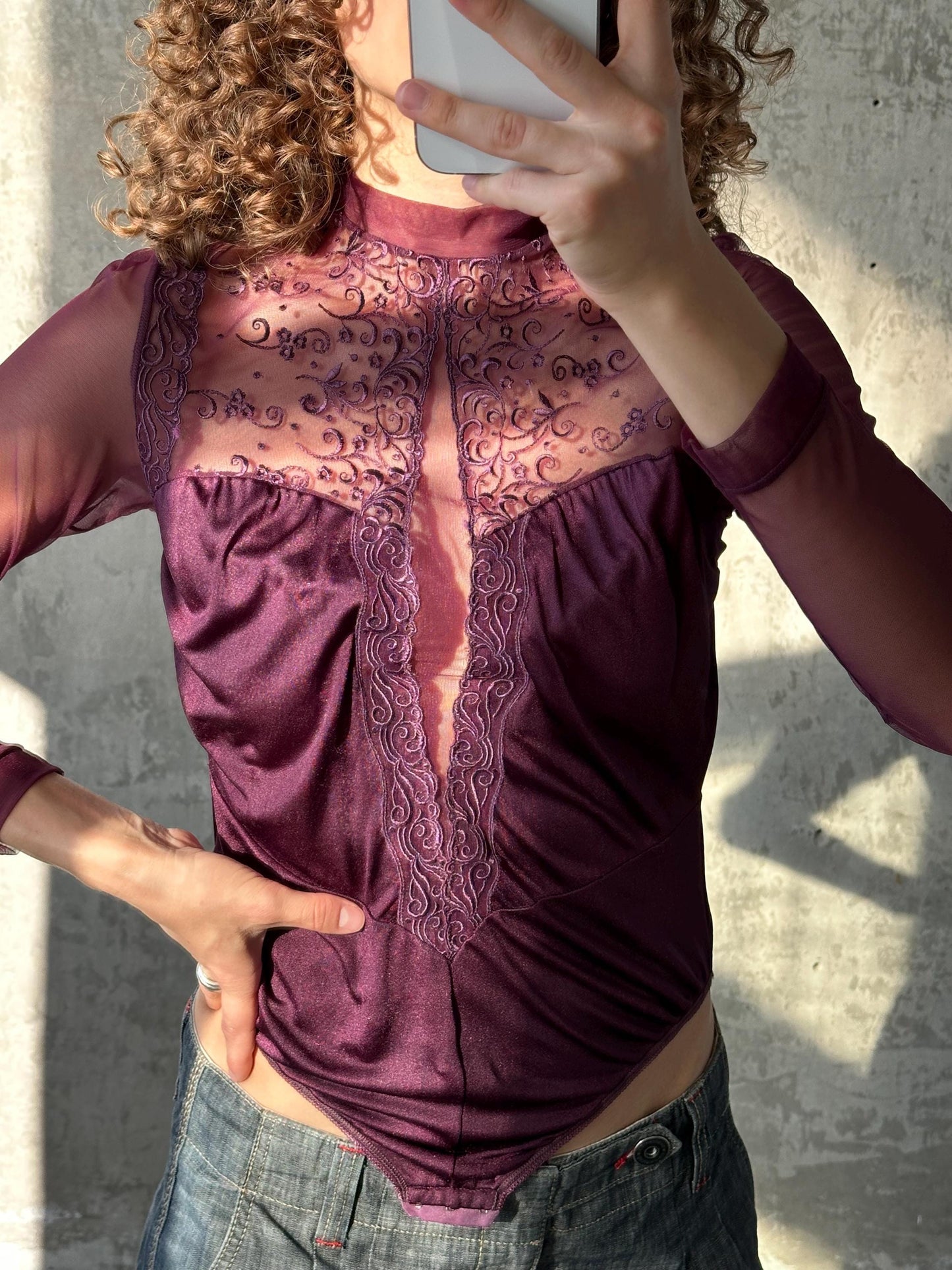 A person is taking a selfie with a smartphone, wearing a purple lace top and blue jeans.