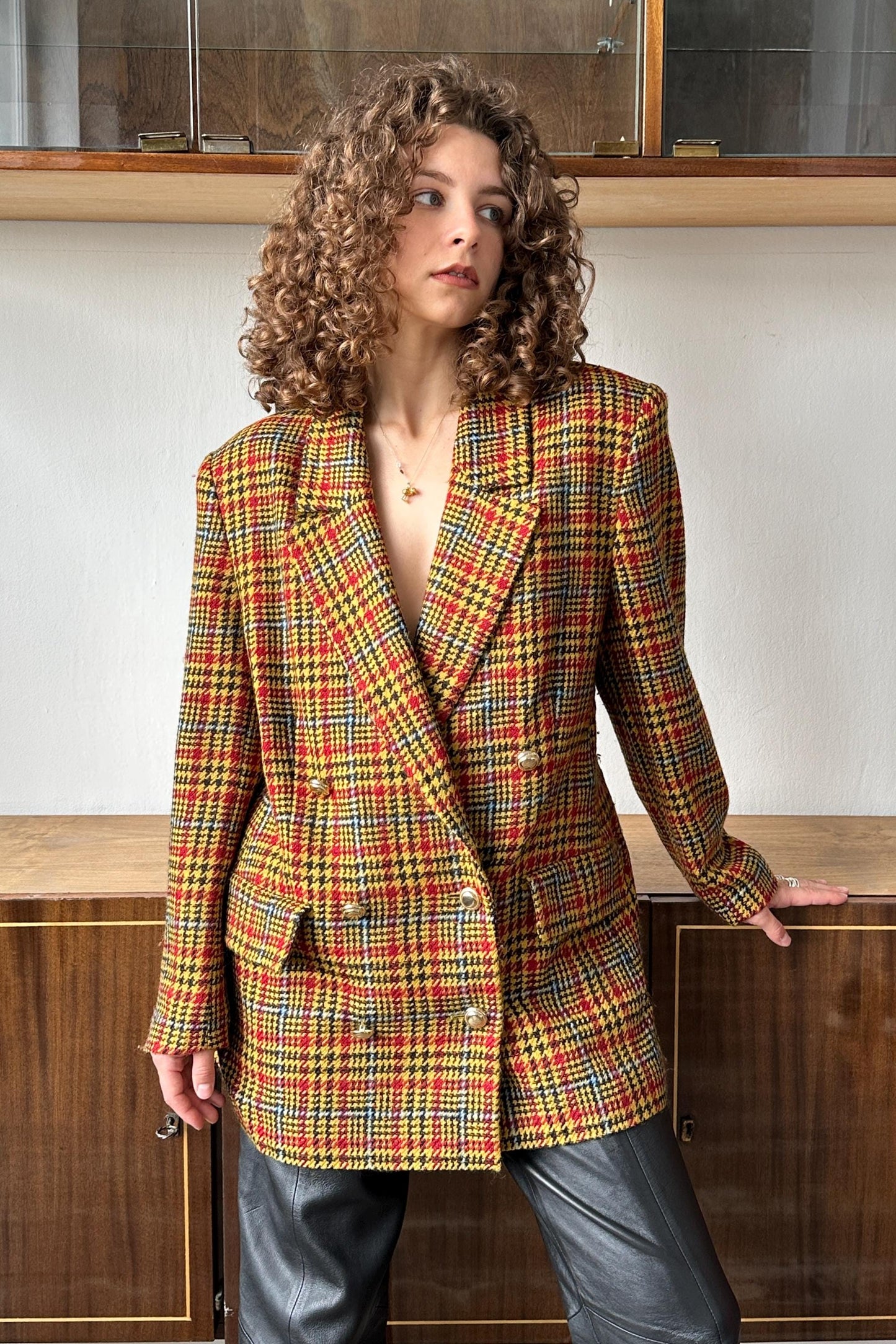 A woman with curly hair wearing a colorful, patterned blazer stands in front of a wooden cabinet.