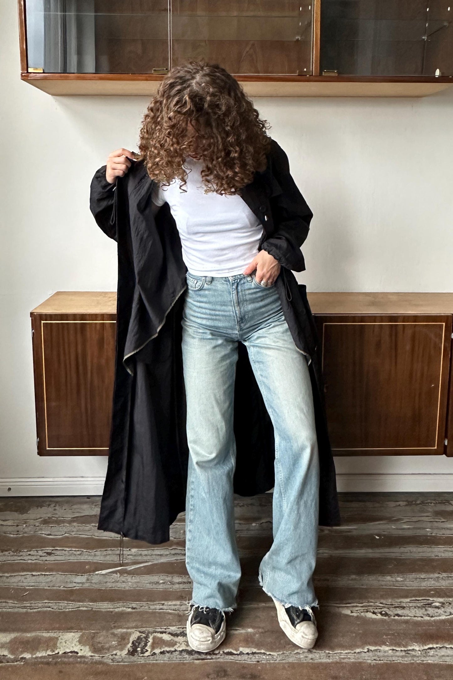 A person with curly hair stands in a room, wearing a black coat, white shirt, and light blue jeans.