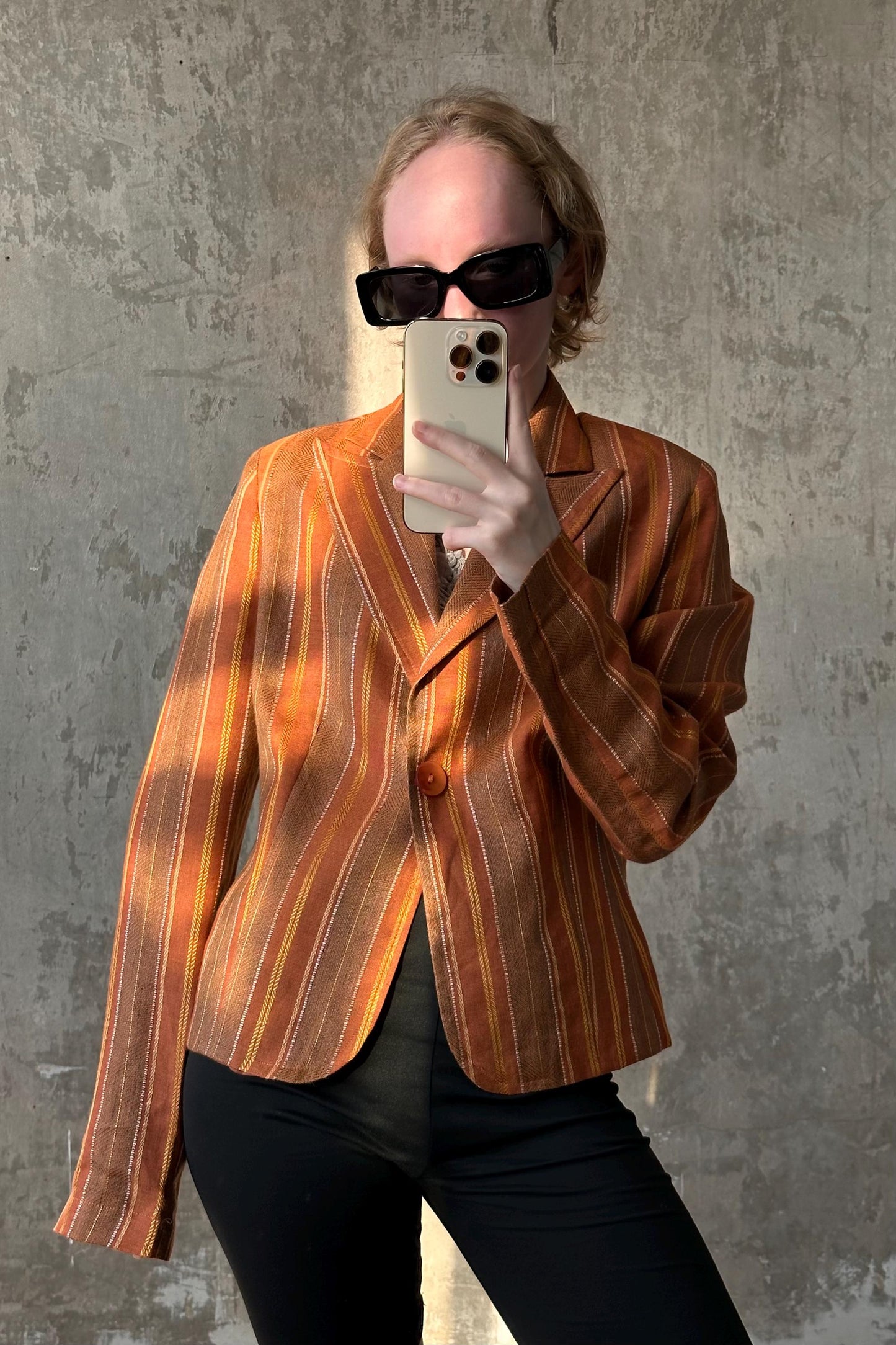 A person wearing a striped orange blazer and sunglasses is taking a selfie with a smartphone.