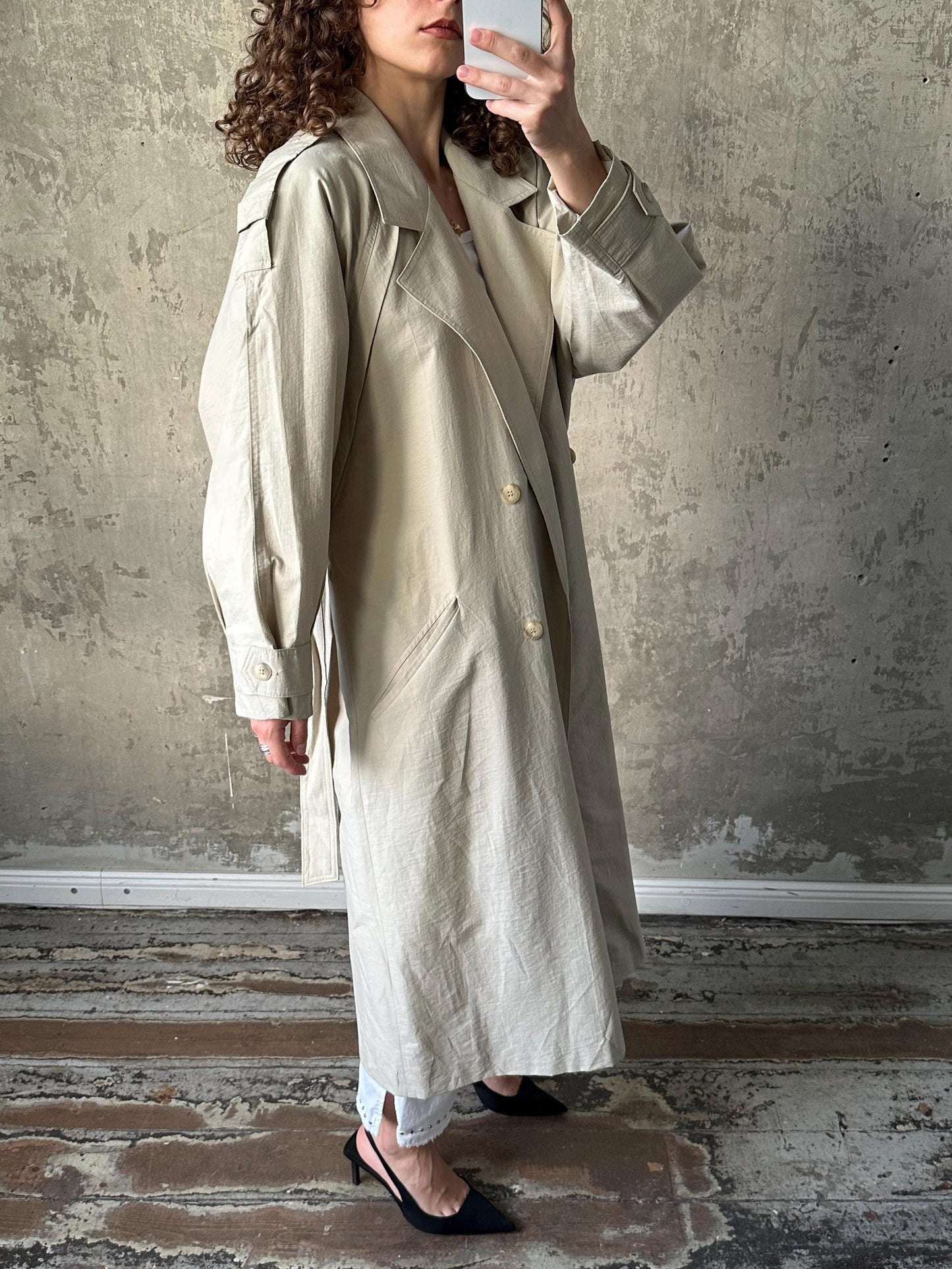 A woman wearing a beige trench coat is taking a selfie with a white phone in front of a concrete wall.