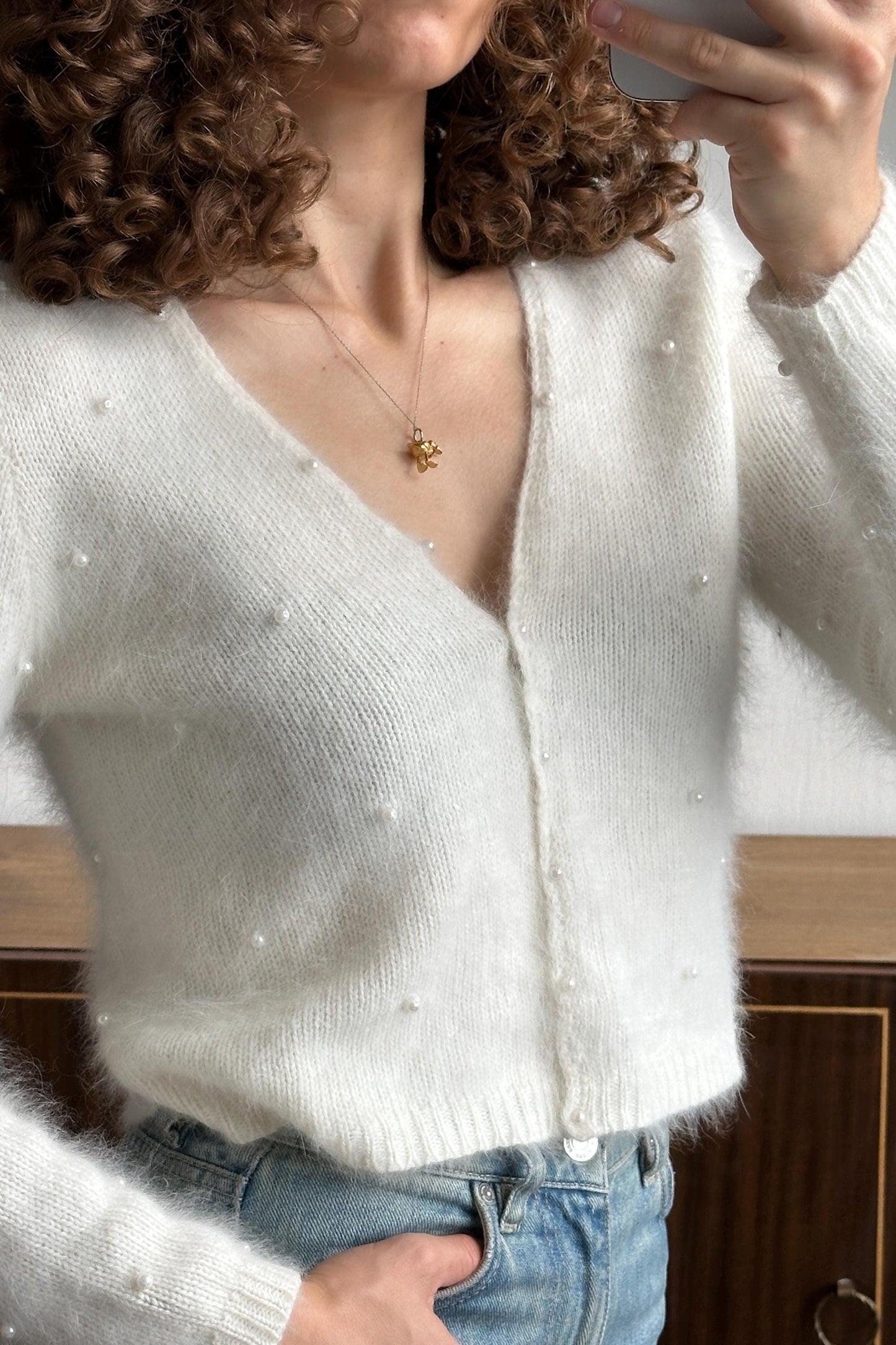 A person with curly hair is taking a selfie with a white cardigan and blue jeans, holding a phone in their hand.
