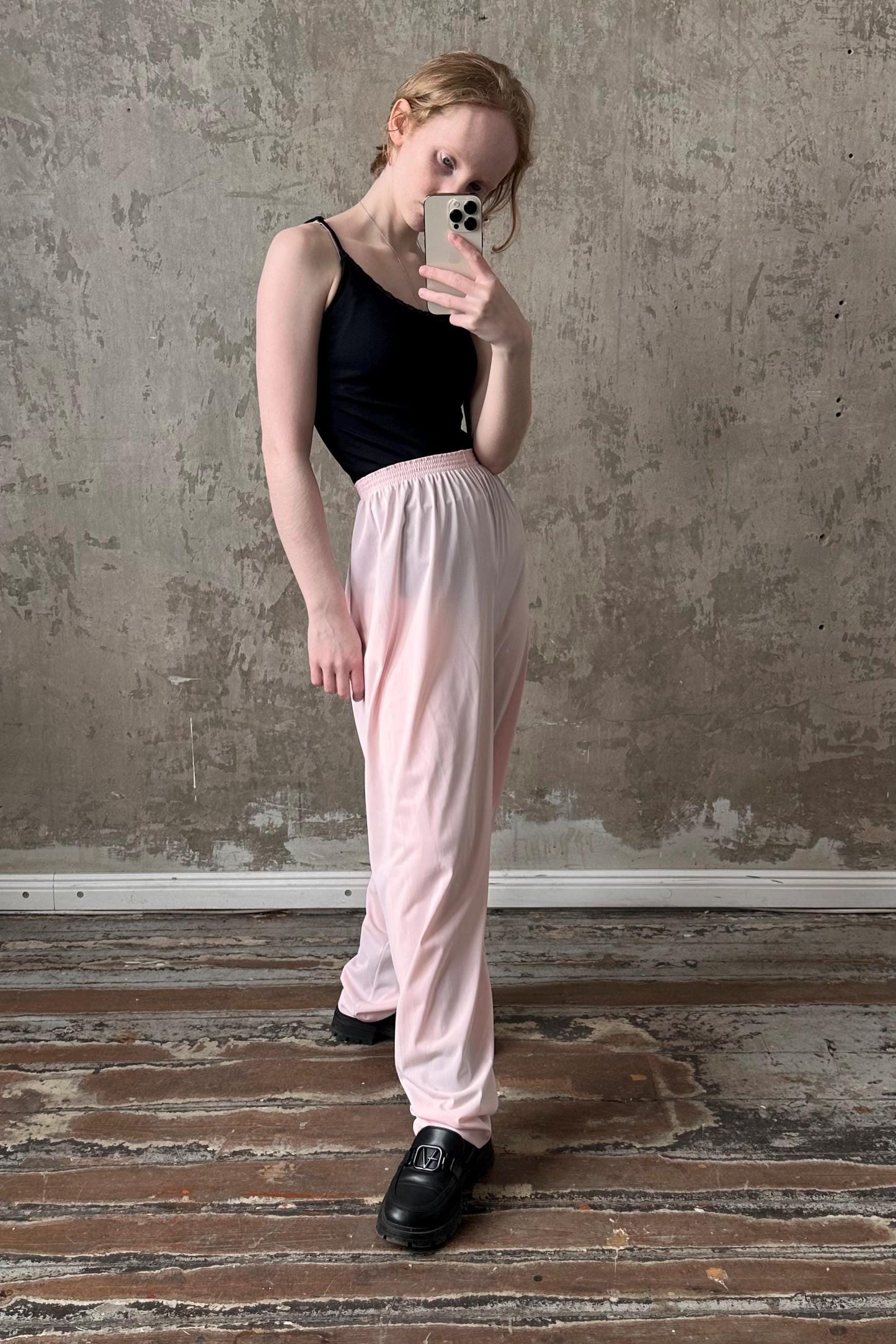 A woman is taking a selfie with a smartphone, wearing a black top and pink pants, standing in front of a concrete wall.