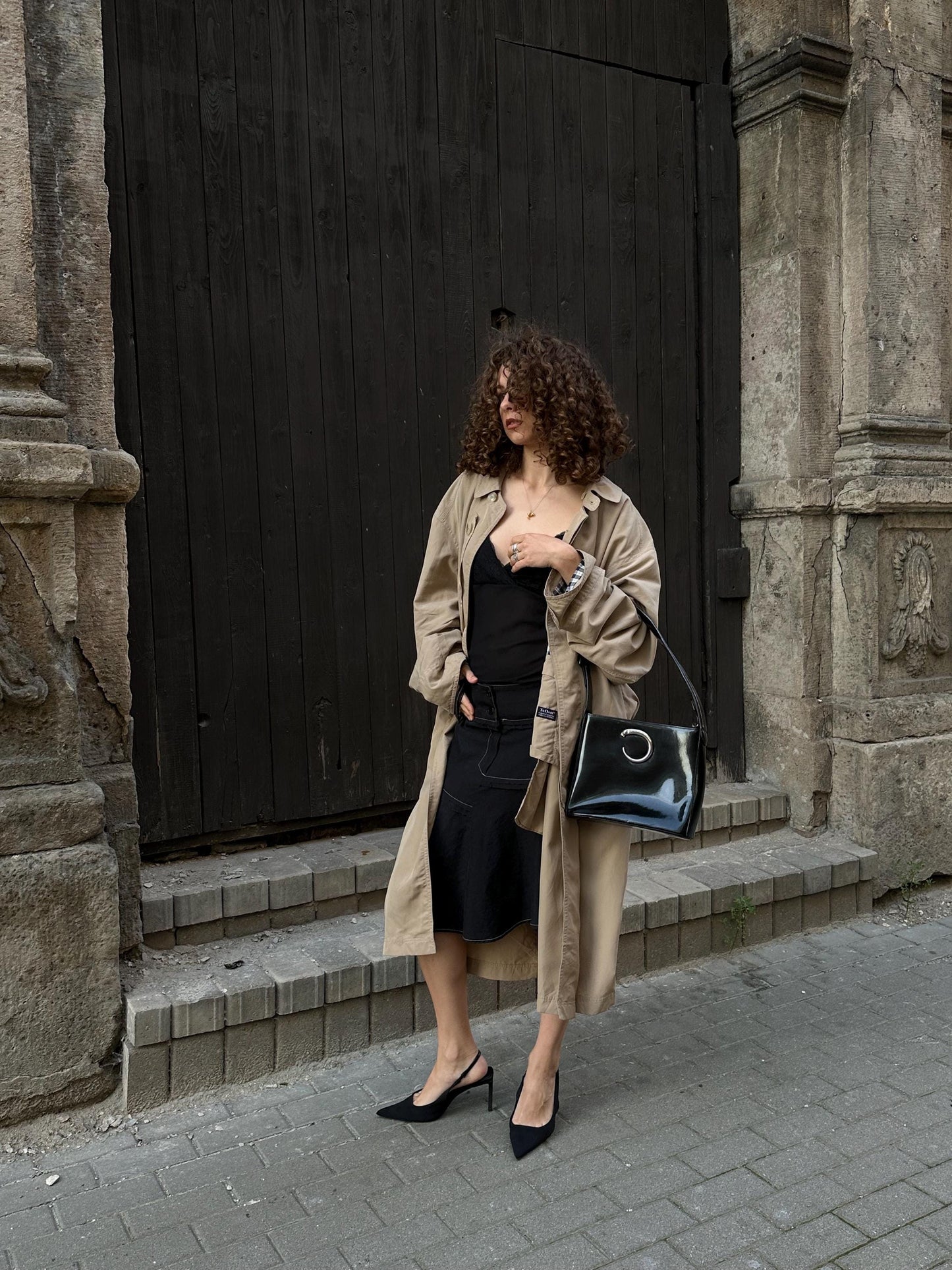 A woman in a beige trench coat and black dress stands in front of a stone building, holding a black purse.