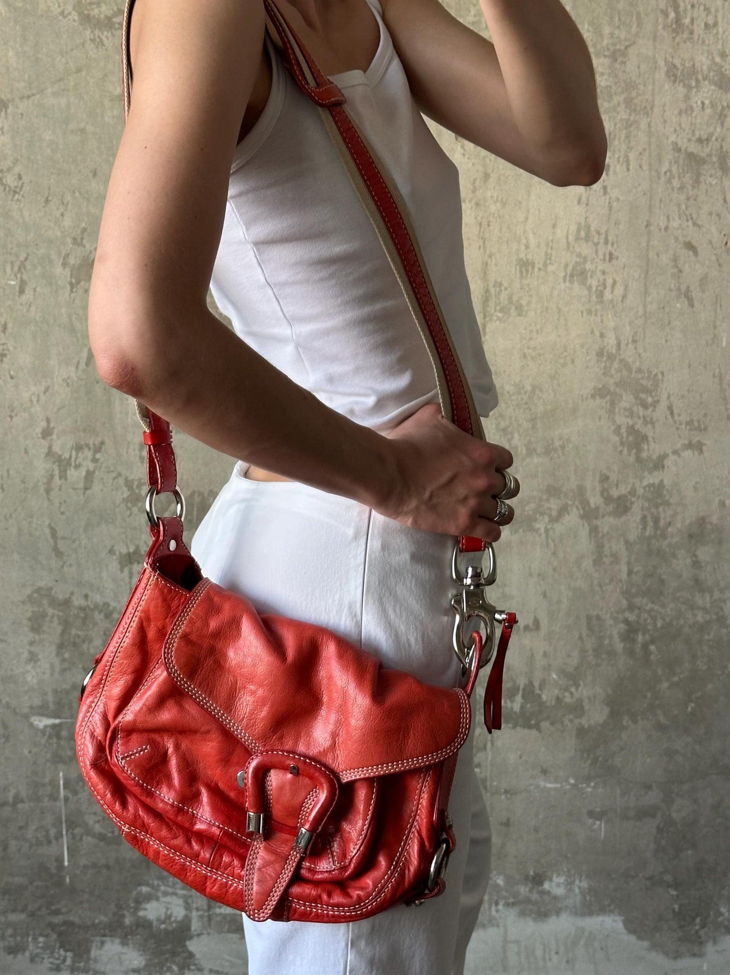 A person wearing a white tank top and white pants is holding a red leather crossbody bag with a red strap.