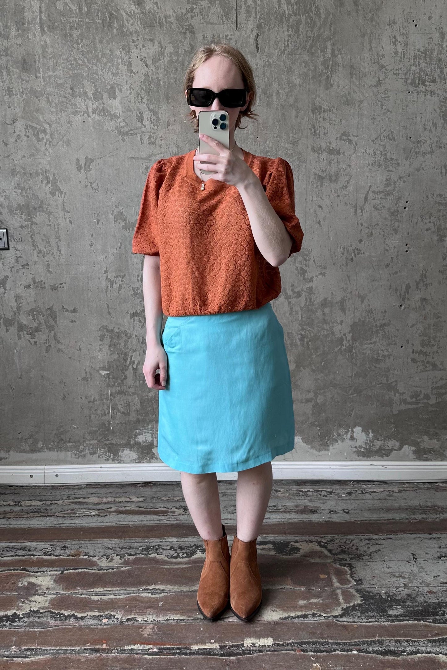 A woman is taking a selfie with a smartphone, wearing an orange top, a blue skirt, and brown boots, standing in front of a concrete wall.