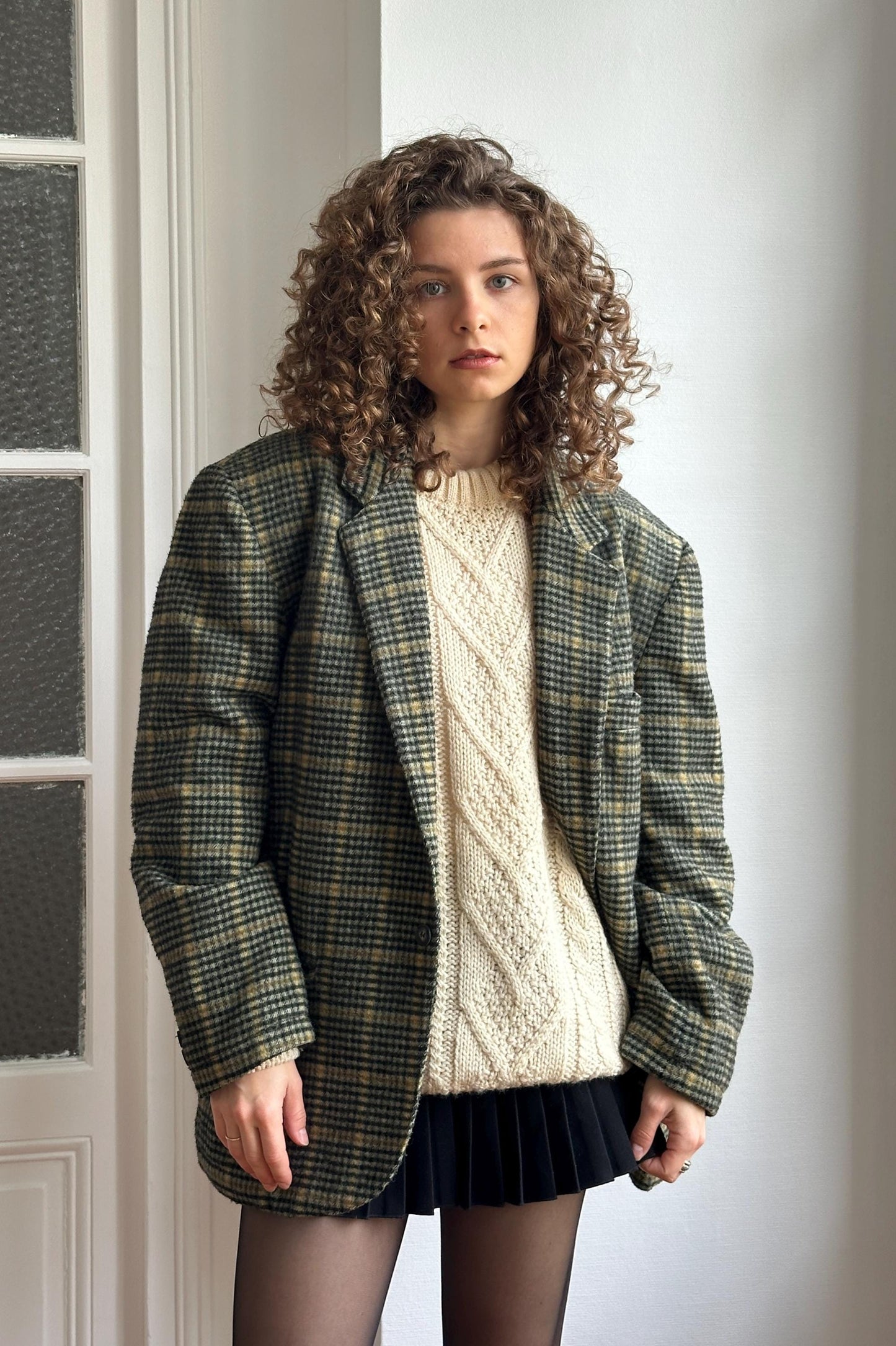 A young woman with curly hair stands in front of a white wall, wearing a plaid jacket, a white sweater, and a black skirt.