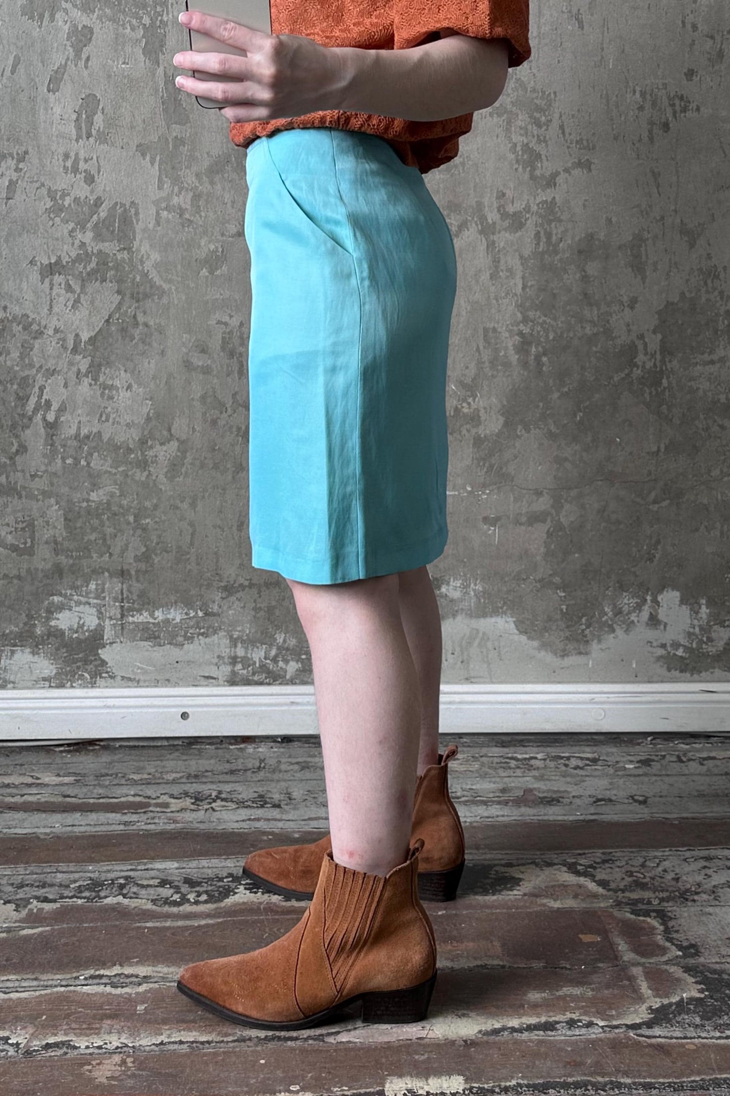 A person wearing a light blue skirt and brown boots stands in front of a concrete wall.