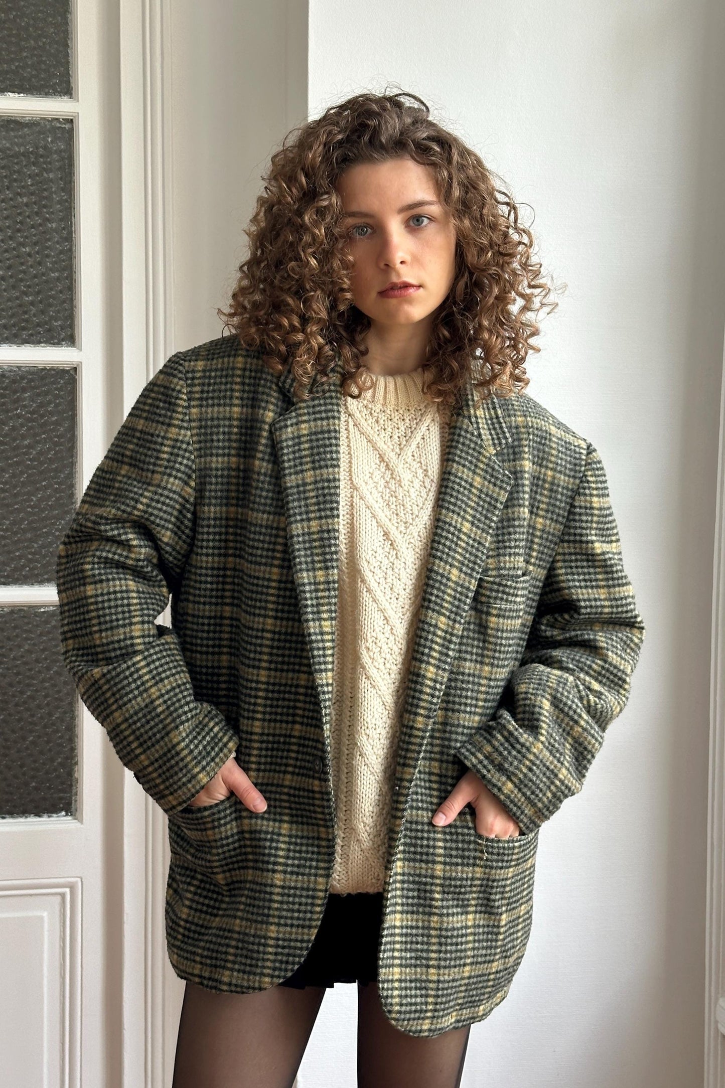 A young woman with curly hair wearing a green and black plaid blazer, a white sweater, and black shorts stands in front of a white wall.