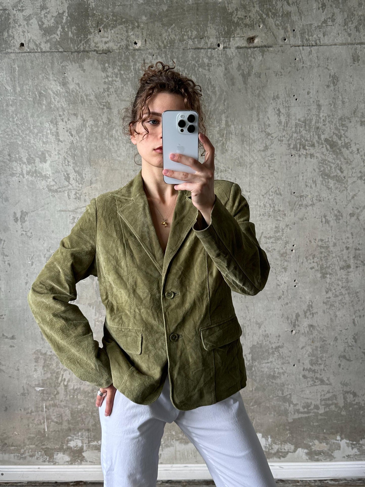A person is taking a selfie with a smartphone, wearing a green blazer and white pants, against a concrete wall background.