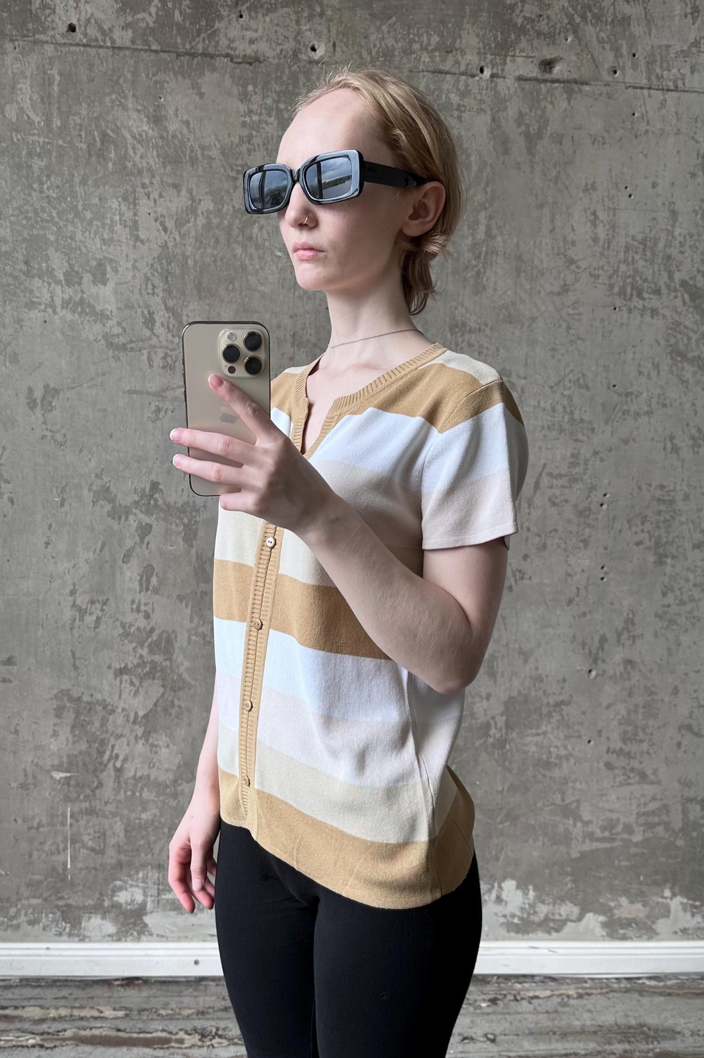 A woman wearing sunglasses and a striped cardigan is taking a selfie with a smartphone against a concrete wall.