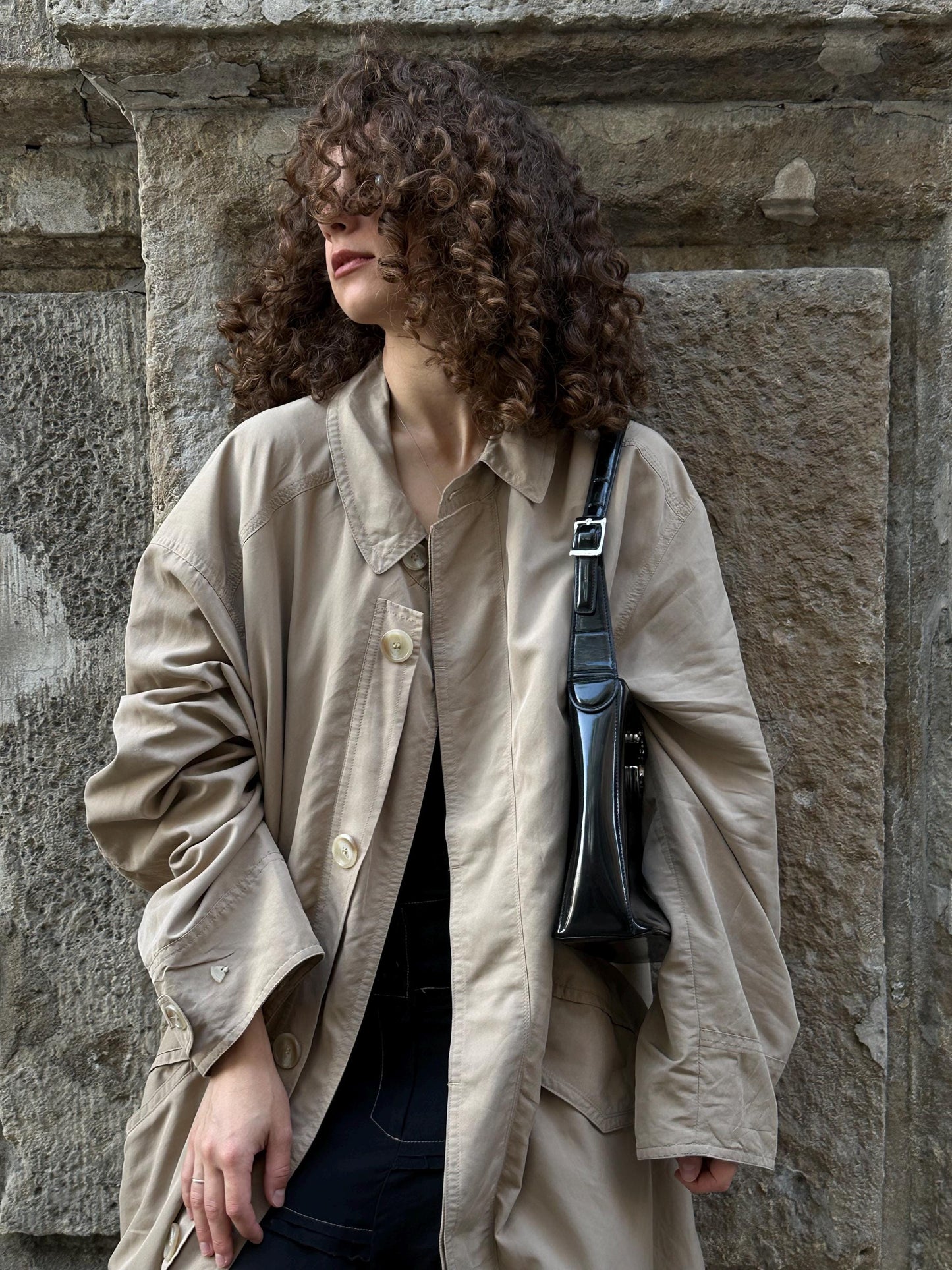 A woman with curly hair stands in front of a stone wall, wearing a beige coat and carrying a black purse.