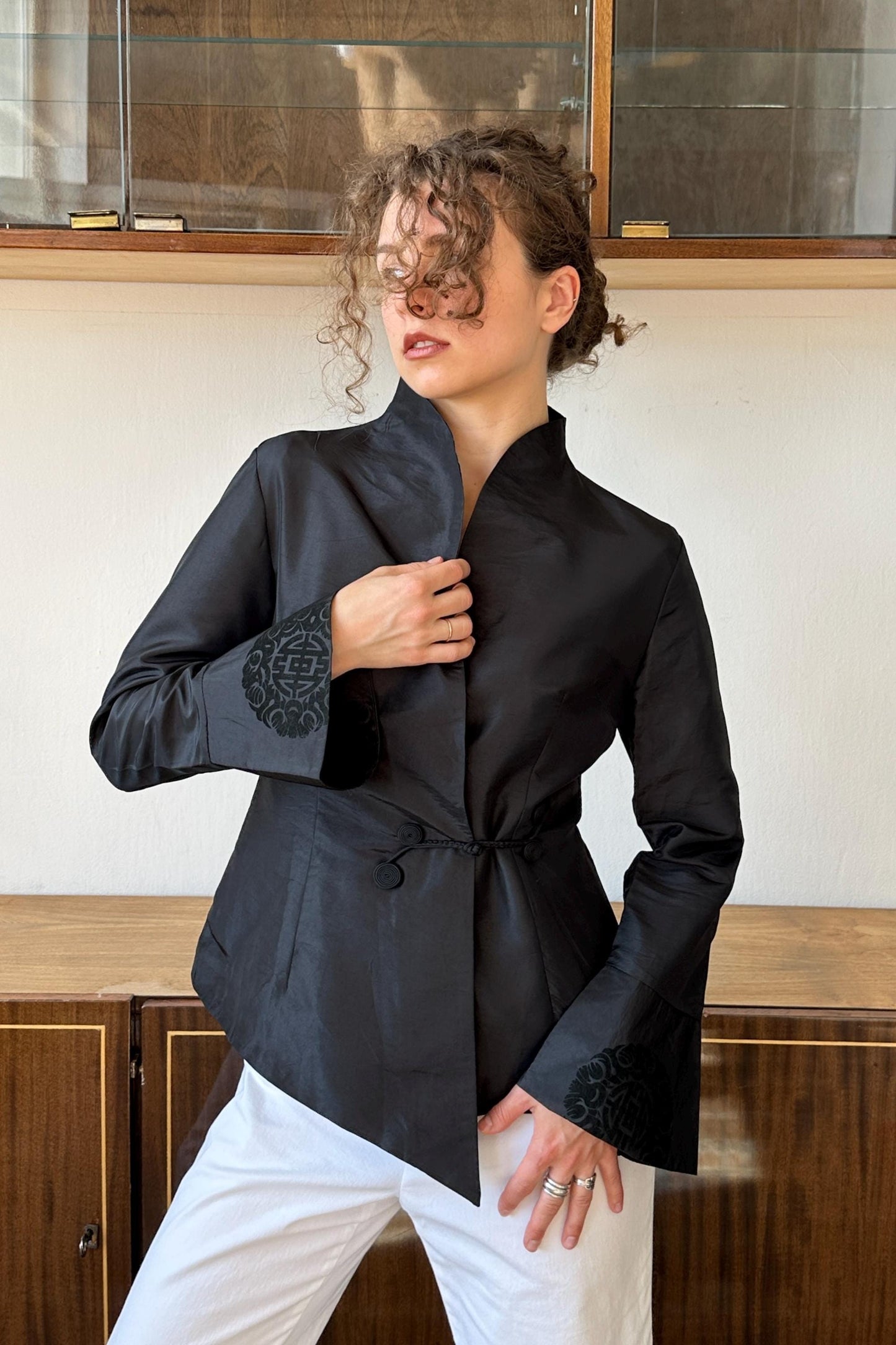 A woman wearing a black jacket with a floral pattern stands in front of a wooden cabinet.