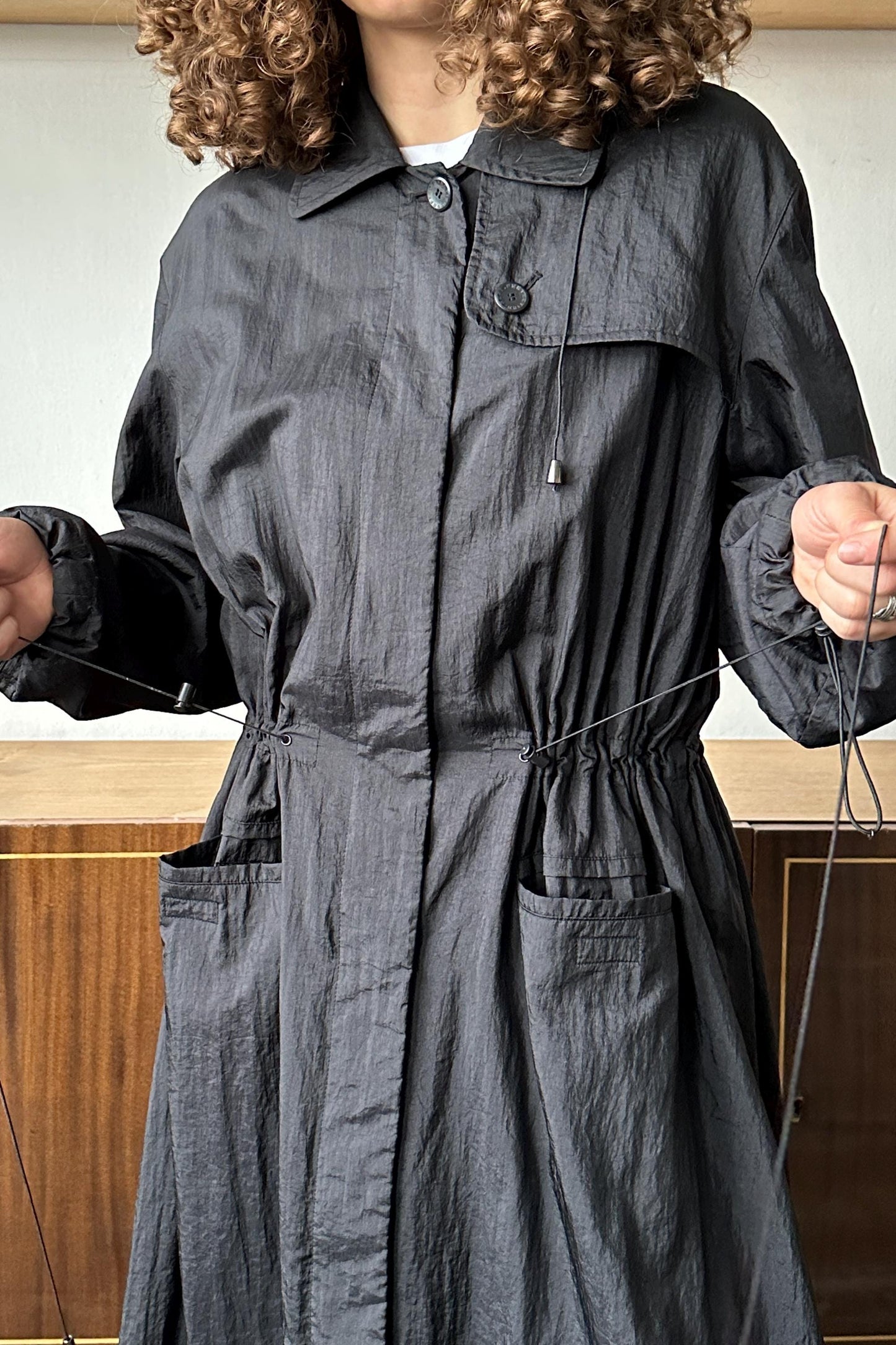 A person wearing a gray coat with curly hair, standing in front of a wooden cabinet.