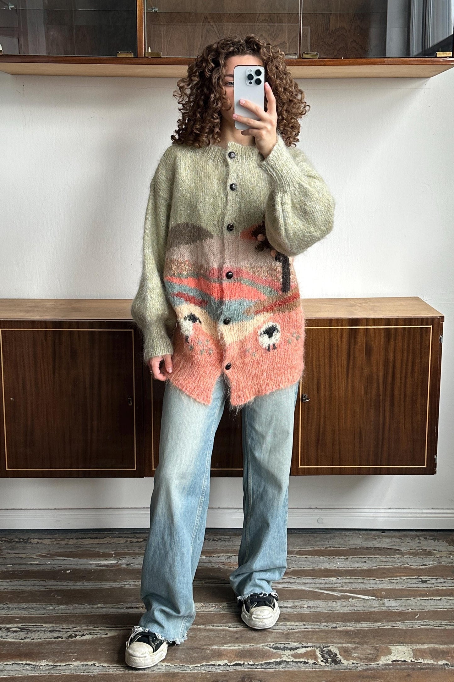 A person with curly hair is taking a selfie with a smartphone, wearing a pink and green knitted cardigan and blue jeans.
