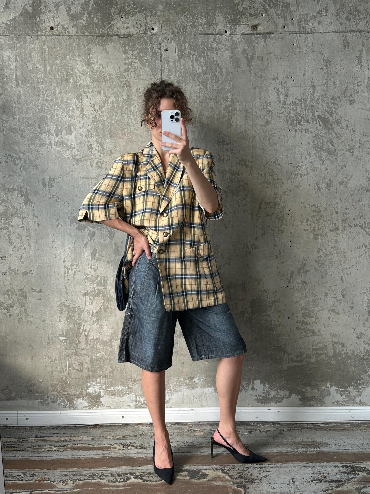 A person is taking a selfie with a smartphone, wearing a plaid jacket, denim shorts, and high heels, against a concrete wall.