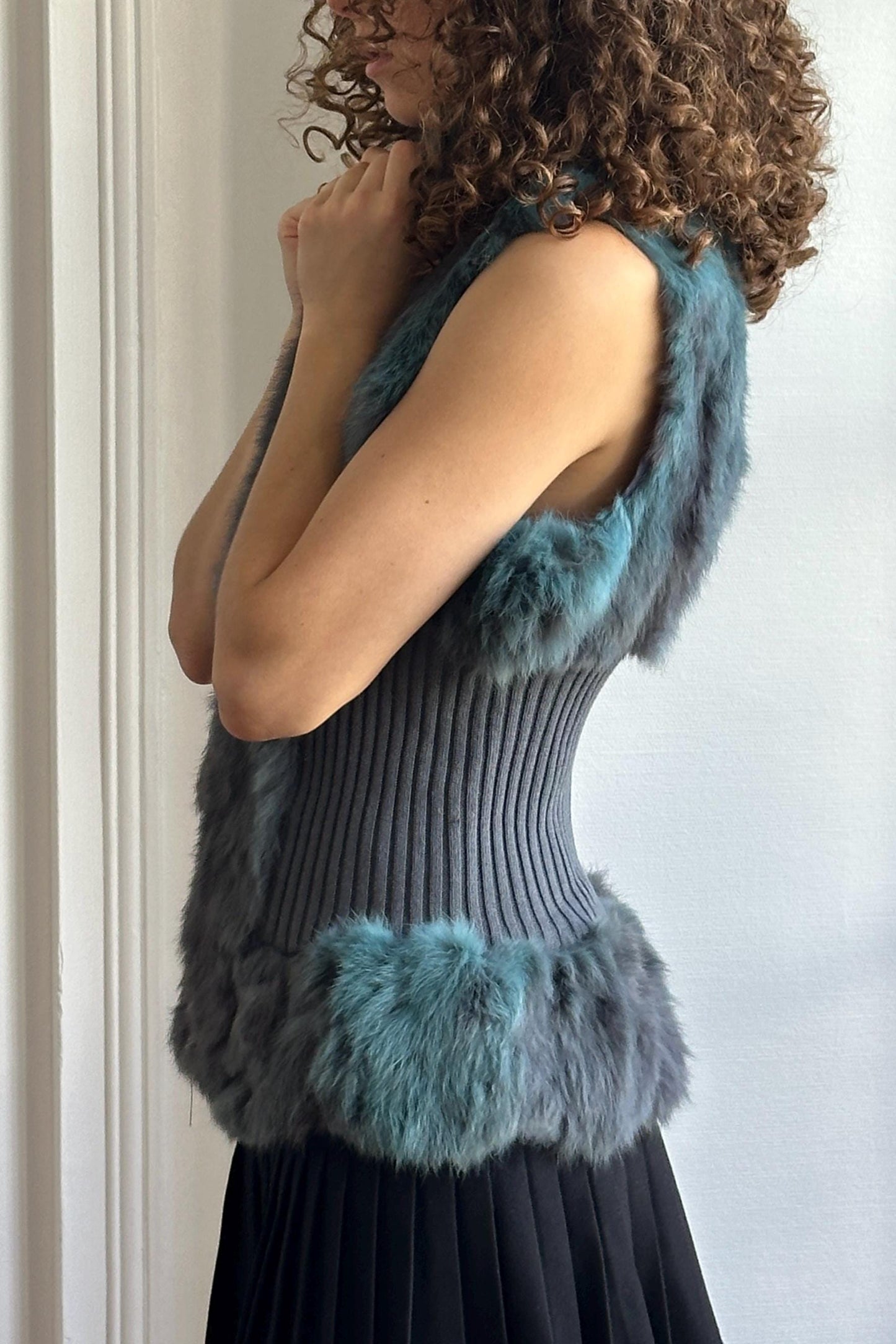 A woman with curly hair wearing a gray and blue fur vest stands in front of a white wall, her hands clasped together.