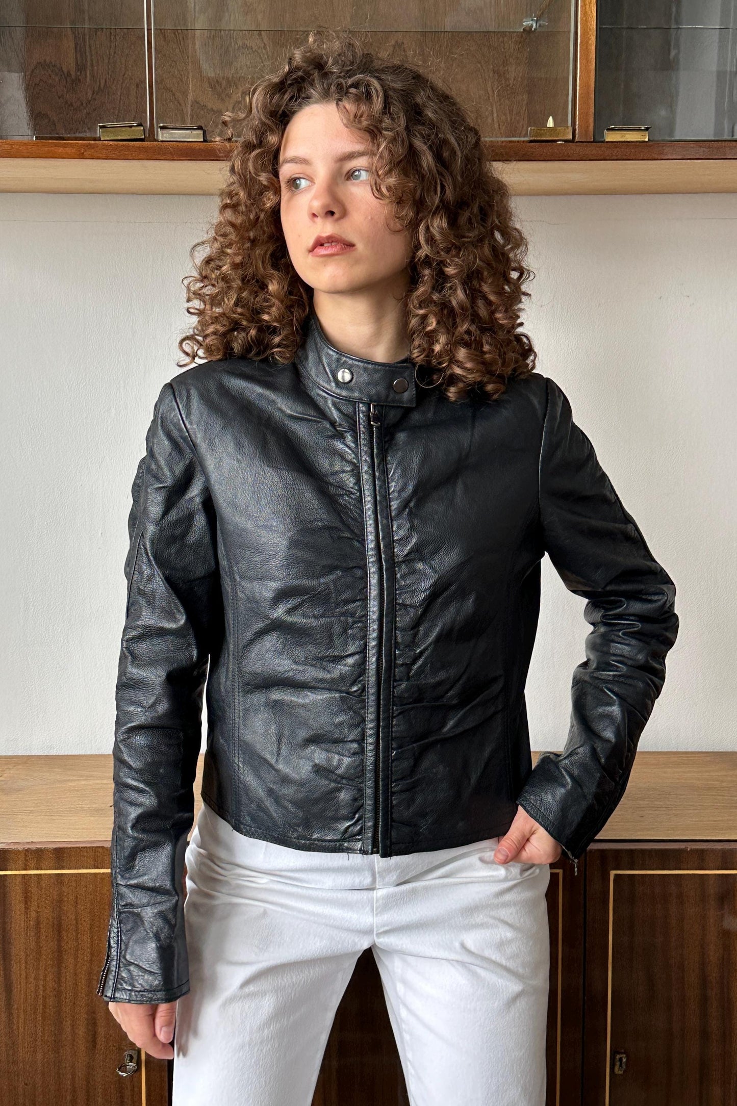 A woman with curly hair wearing a black leather jacket stands in front of a wooden cabinet.