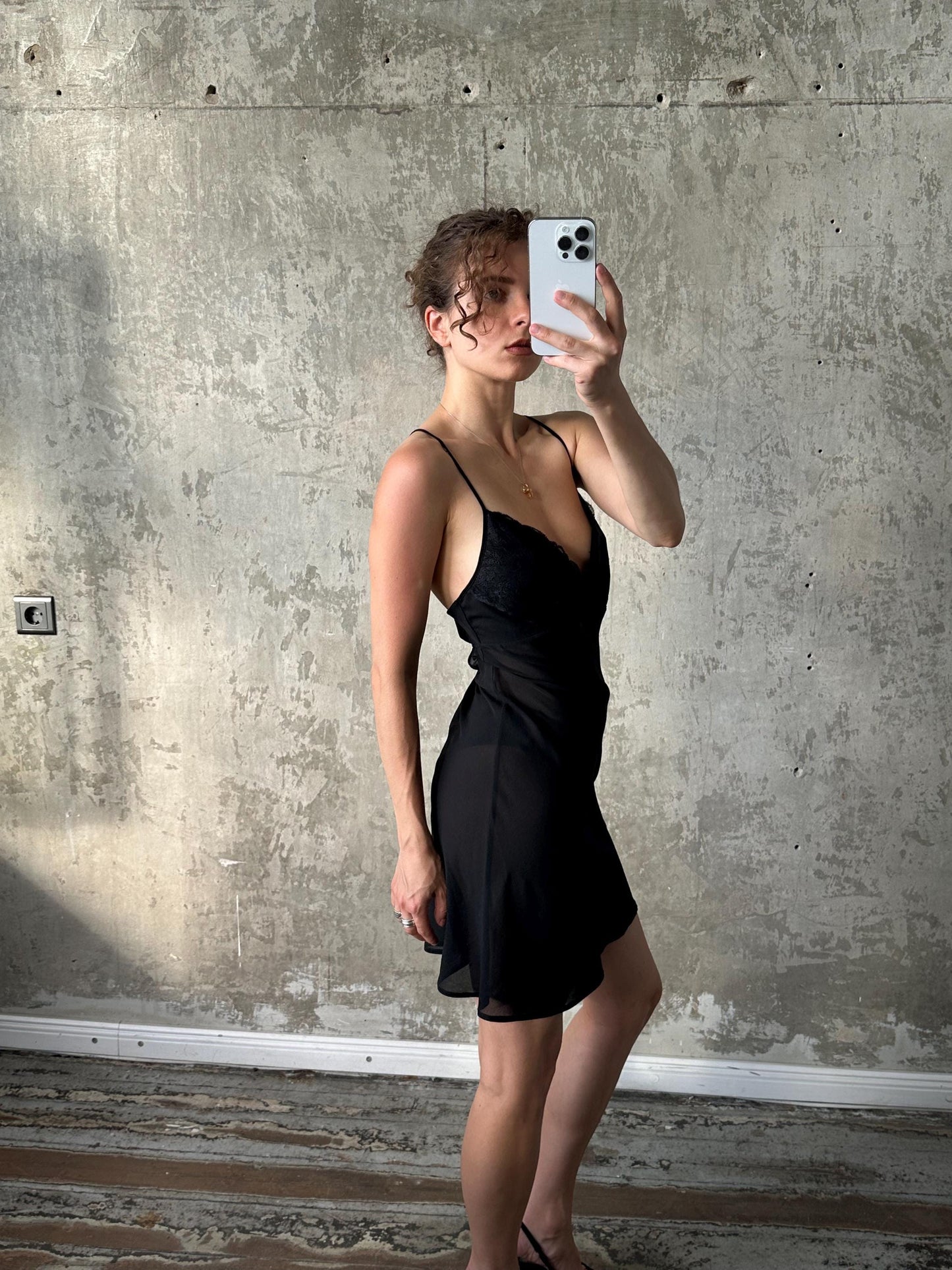A woman in a black dress is taking a selfie with a white iPhone in front of a concrete wall.