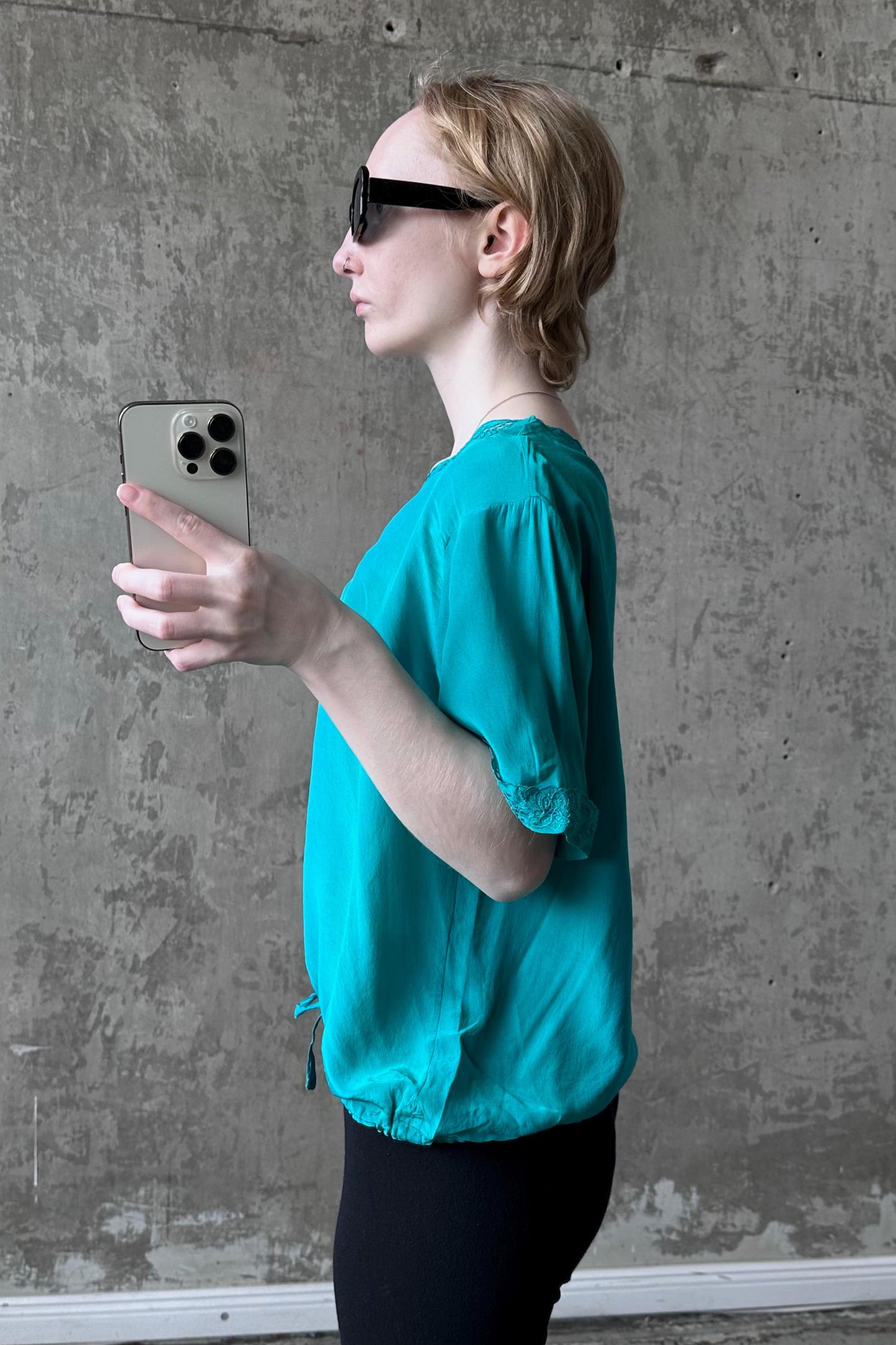 A woman in a teal blouse and sunglasses is holding a smartphone and looking at it, standing in front of a concrete wall.