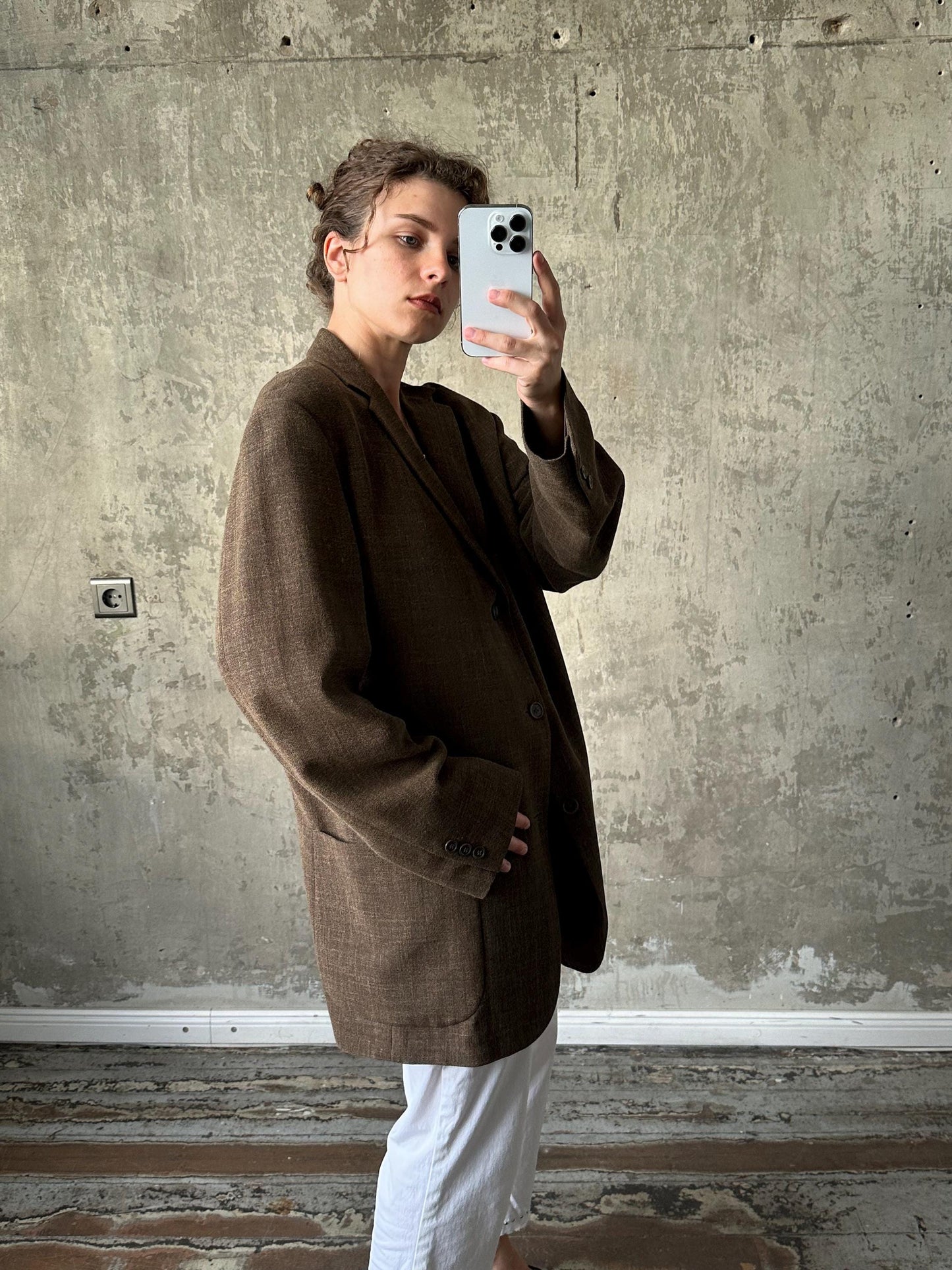 A person is taking a selfie with a smartphone, wearing a brown blazer and white pants, against a concrete wall background.