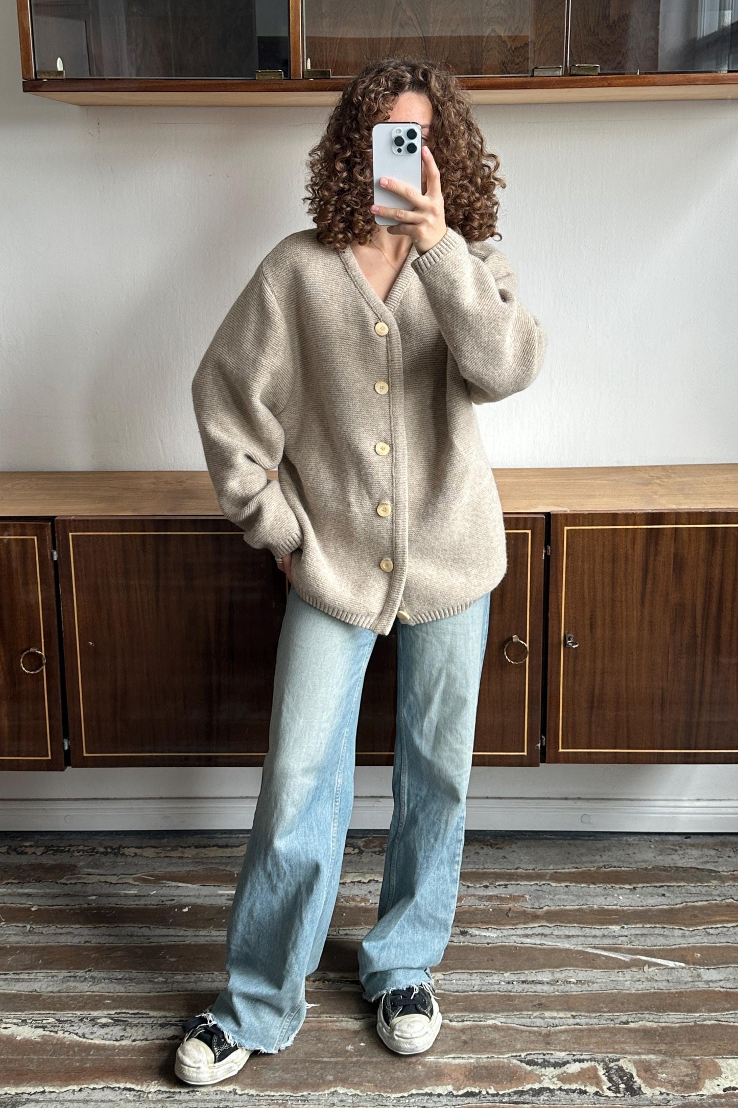 A person with curly hair is taking a selfie with a smartphone, standing in front of a wooden cabinet.