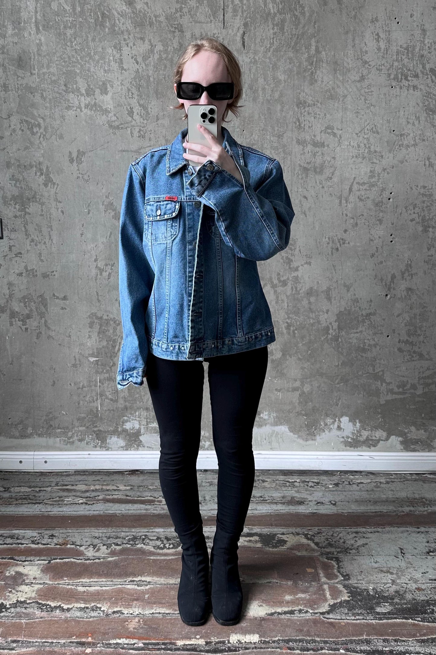 A person wearing a denim jacket, black leggings, and black boots is taking a selfie with a smartphone in front of a concrete wall.