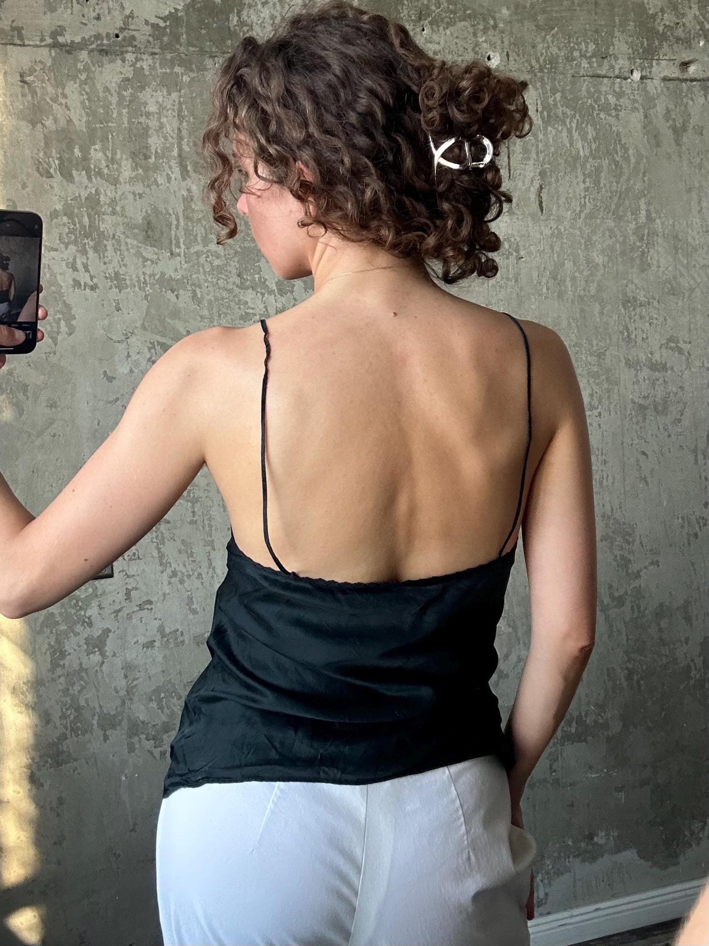 a woman taking a selfie in front of a concrete wall. She is wearing a black top and white pants, and her hair is styled in a bun.
