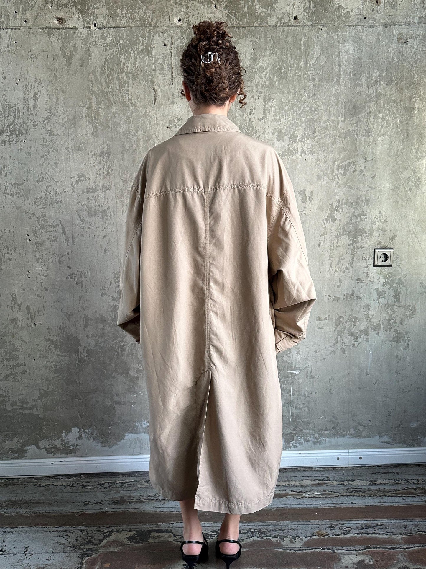 A person wearing a beige coat stands in front of a concrete wall, facing away from the camera.