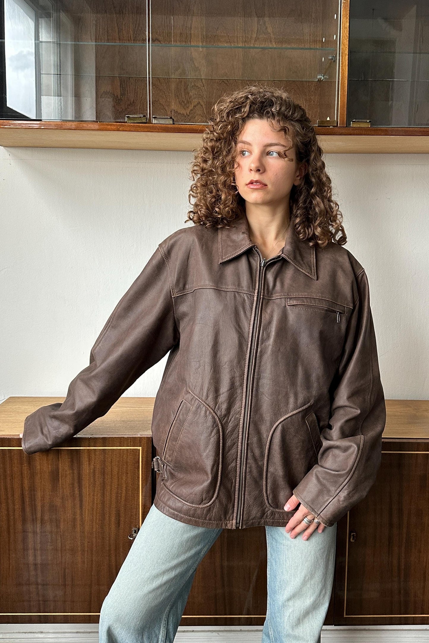 A woman with curly hair wearing a brown leather jacket stands in front of a wooden cabinet.