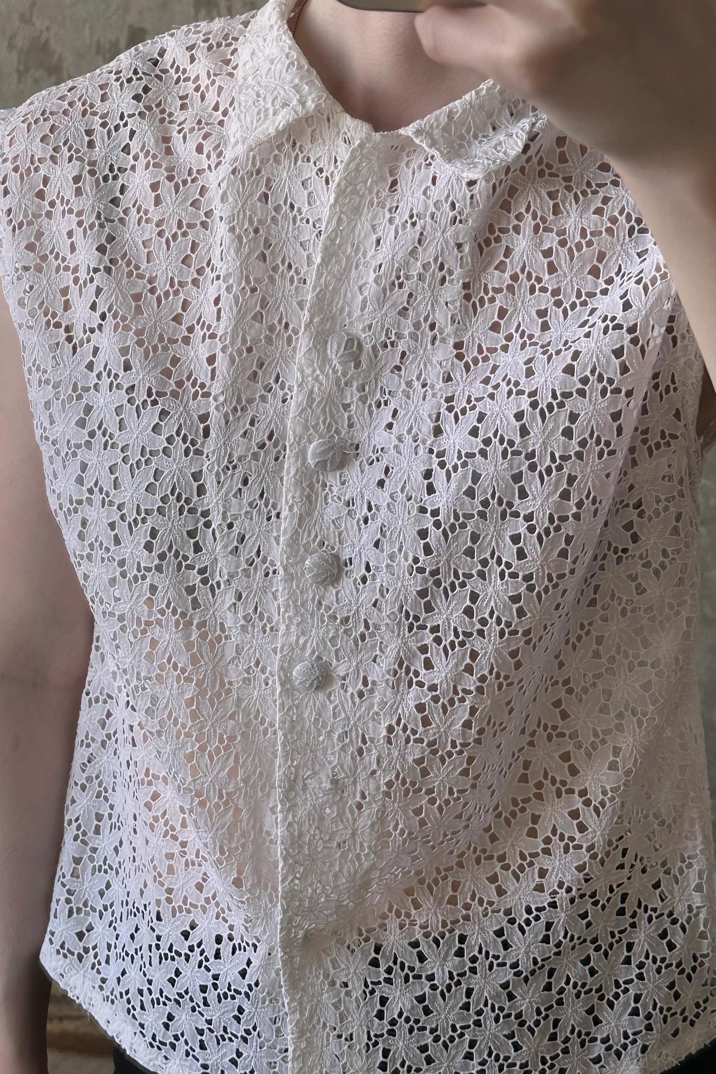 A close-up view of a white lace shirt with a floral pattern, featuring a button-down front and a sleeveless design.