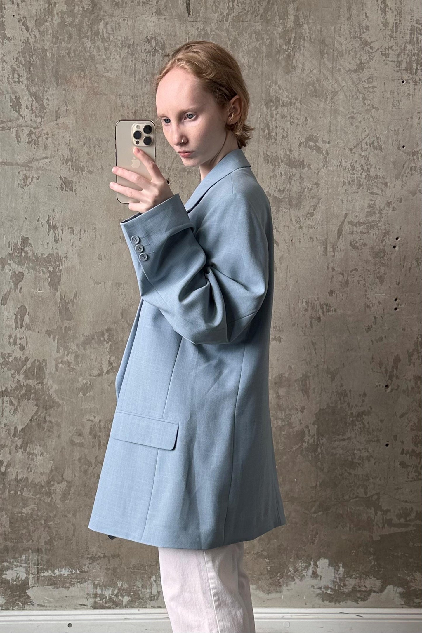 A person wearing a light blue blazer is taking a selfie with a smartphone against a textured concrete wall.