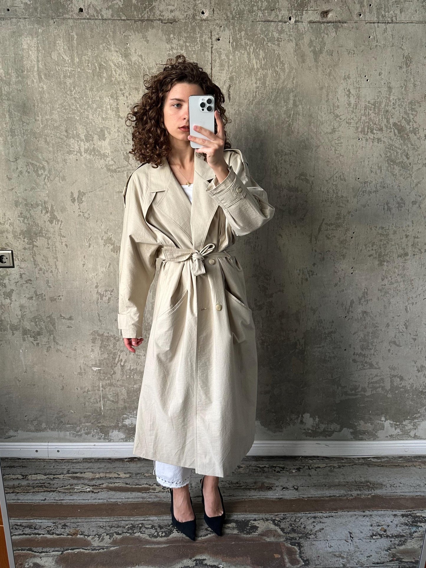 A woman in a beige trench coat is taking a selfie with a white phone against a concrete wall.