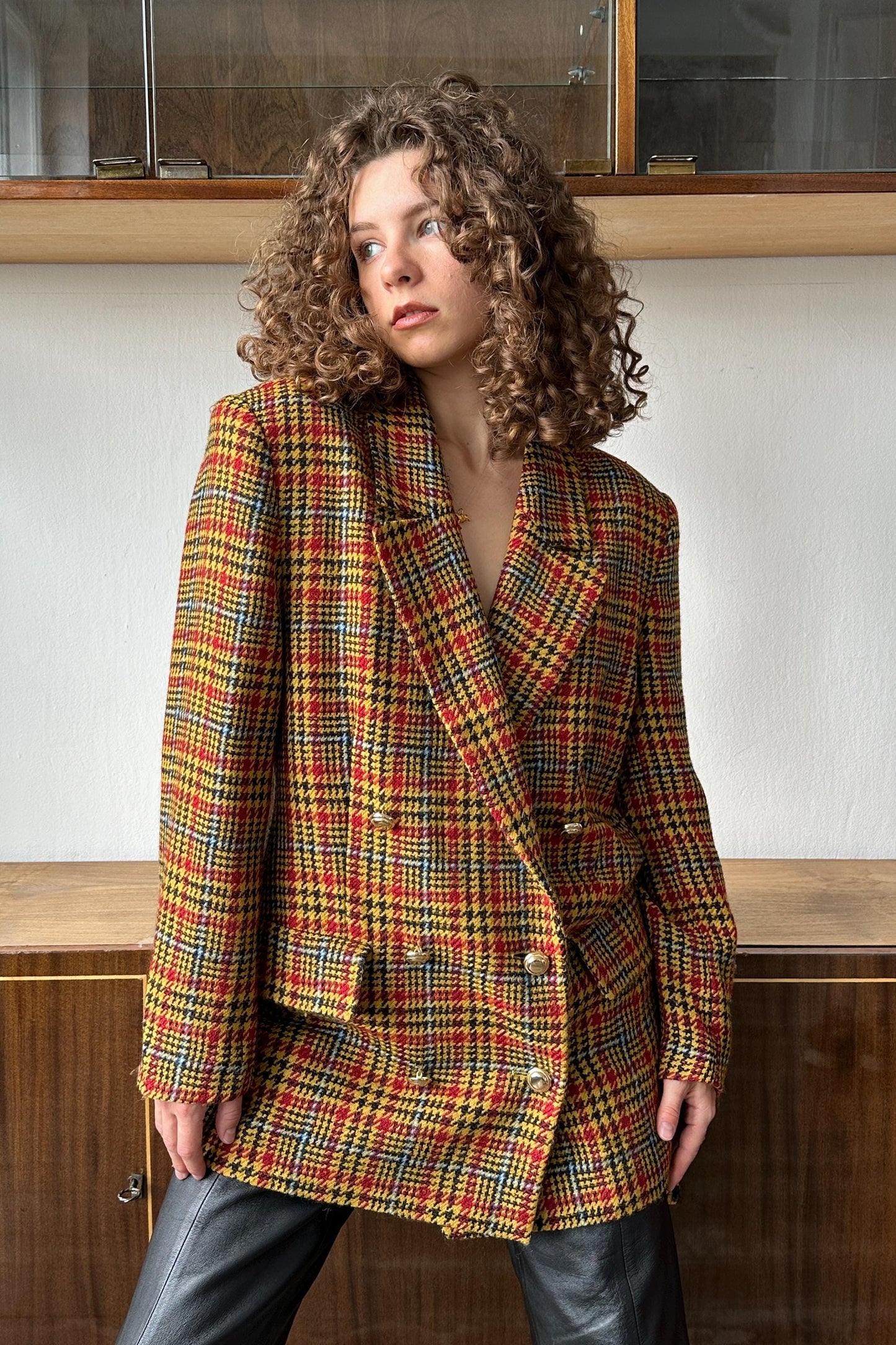 A woman with curly hair wearing a colorful, patterned coat stands in front of a wooden cabinet.