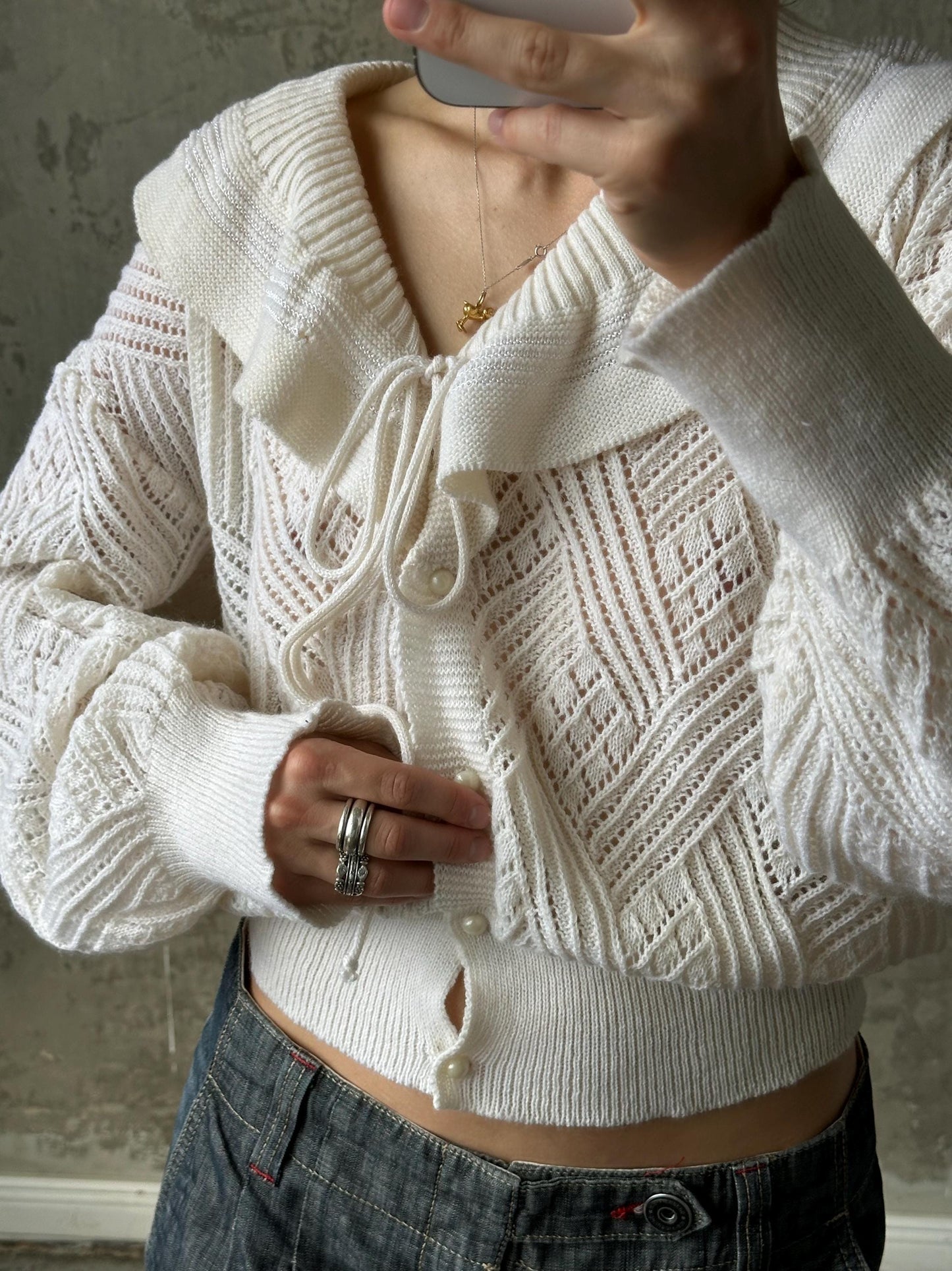 A person is taking a selfie with a white knitted cardigan and jeans, holding a phone up to their face.