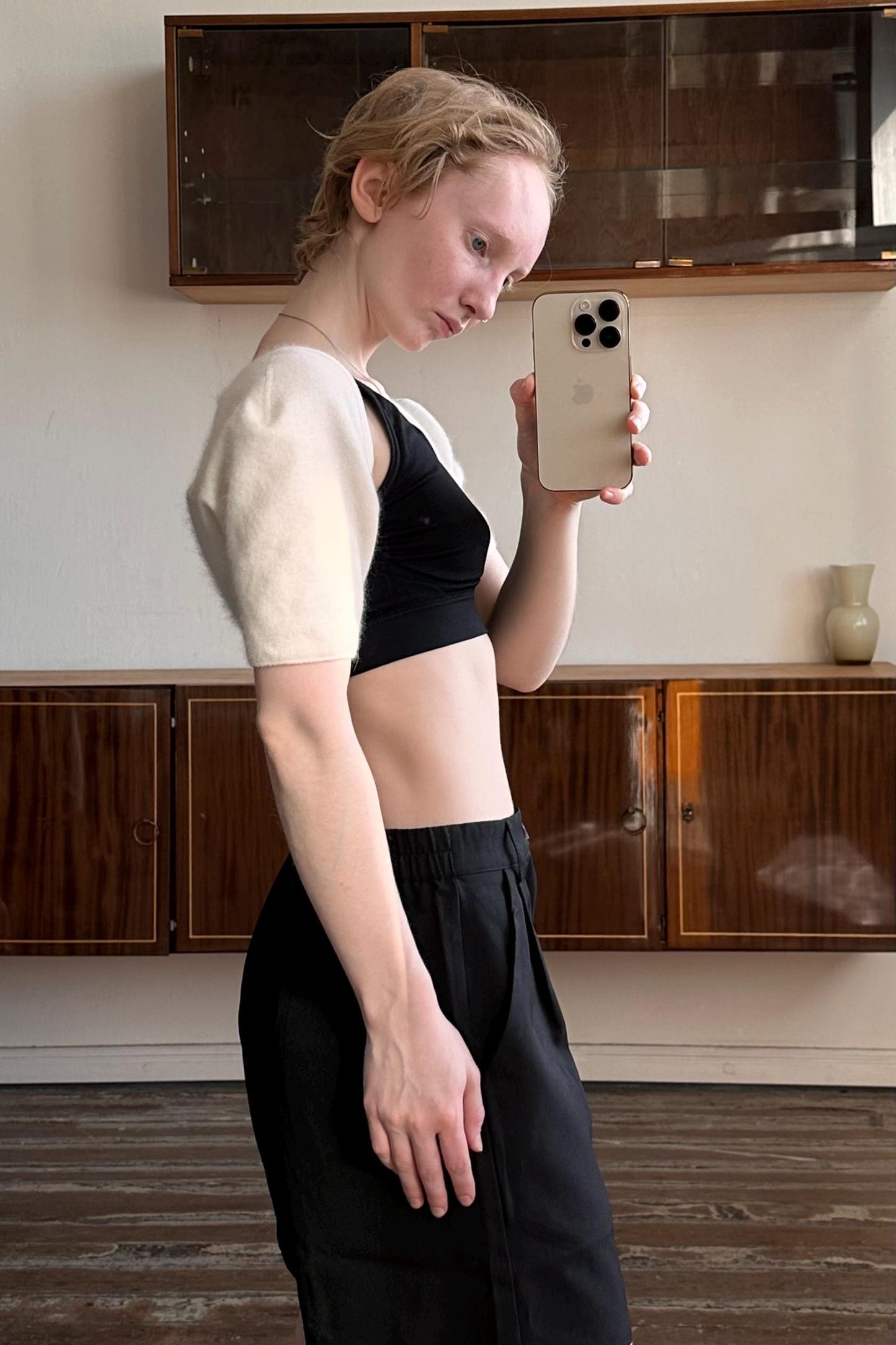 A woman in a black top and black pants is taking a selfie with a white iPhone in a room with wooden cabinets.