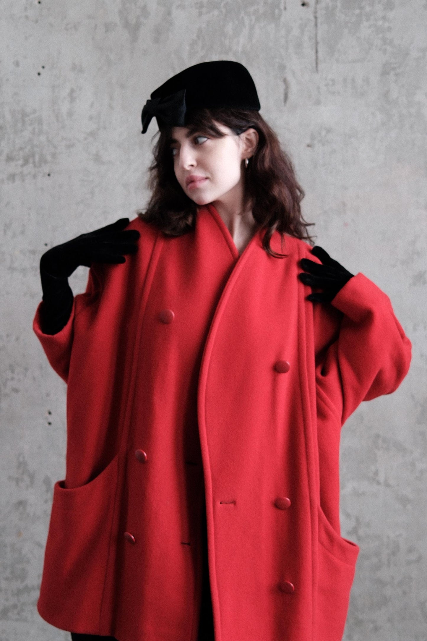 A woman wearing a red coat and black gloves stands against a gray background.
