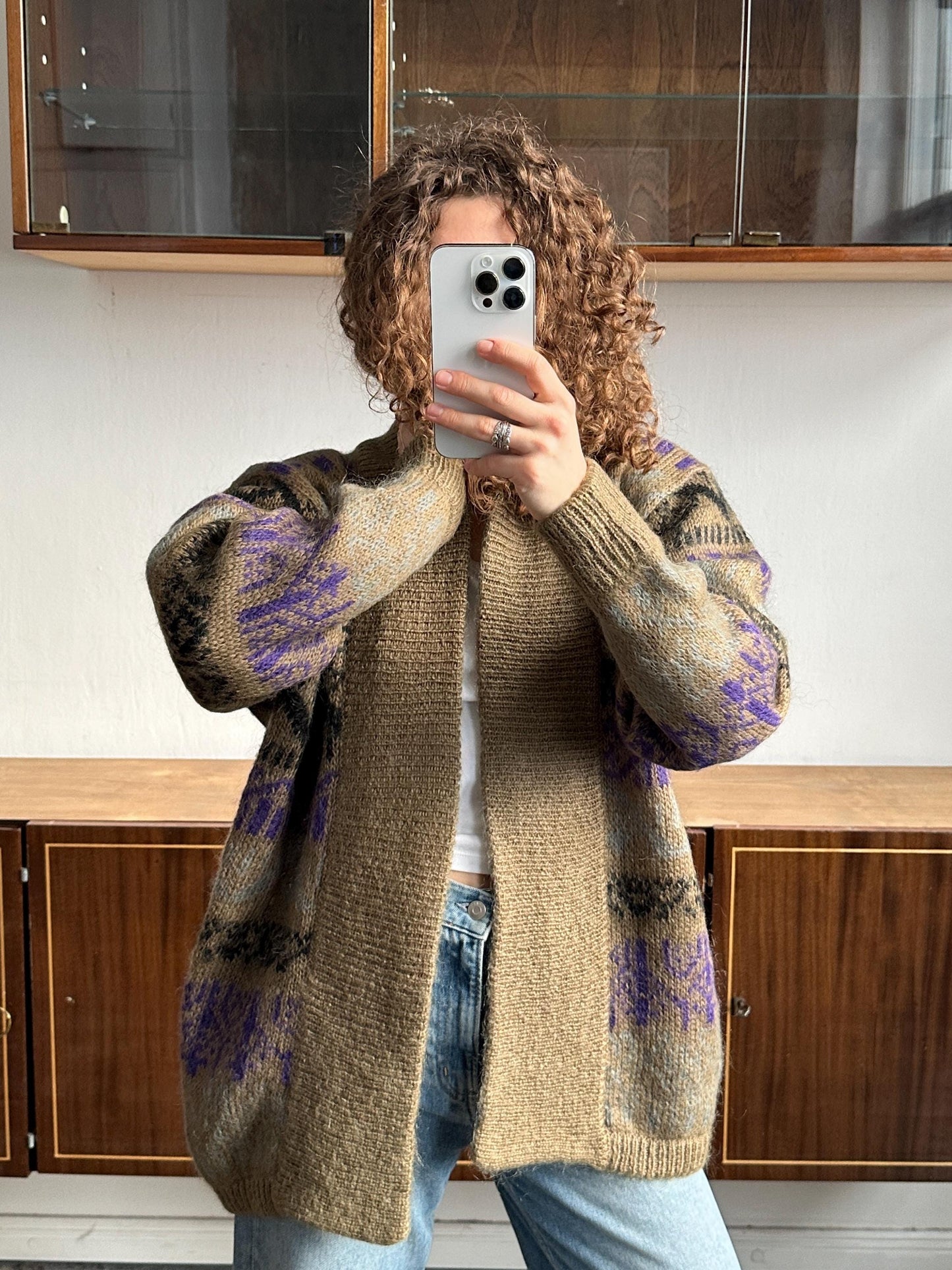 A person with curly hair is taking a selfie with a smartphone, wearing a beige sweater with purple and black patterns.