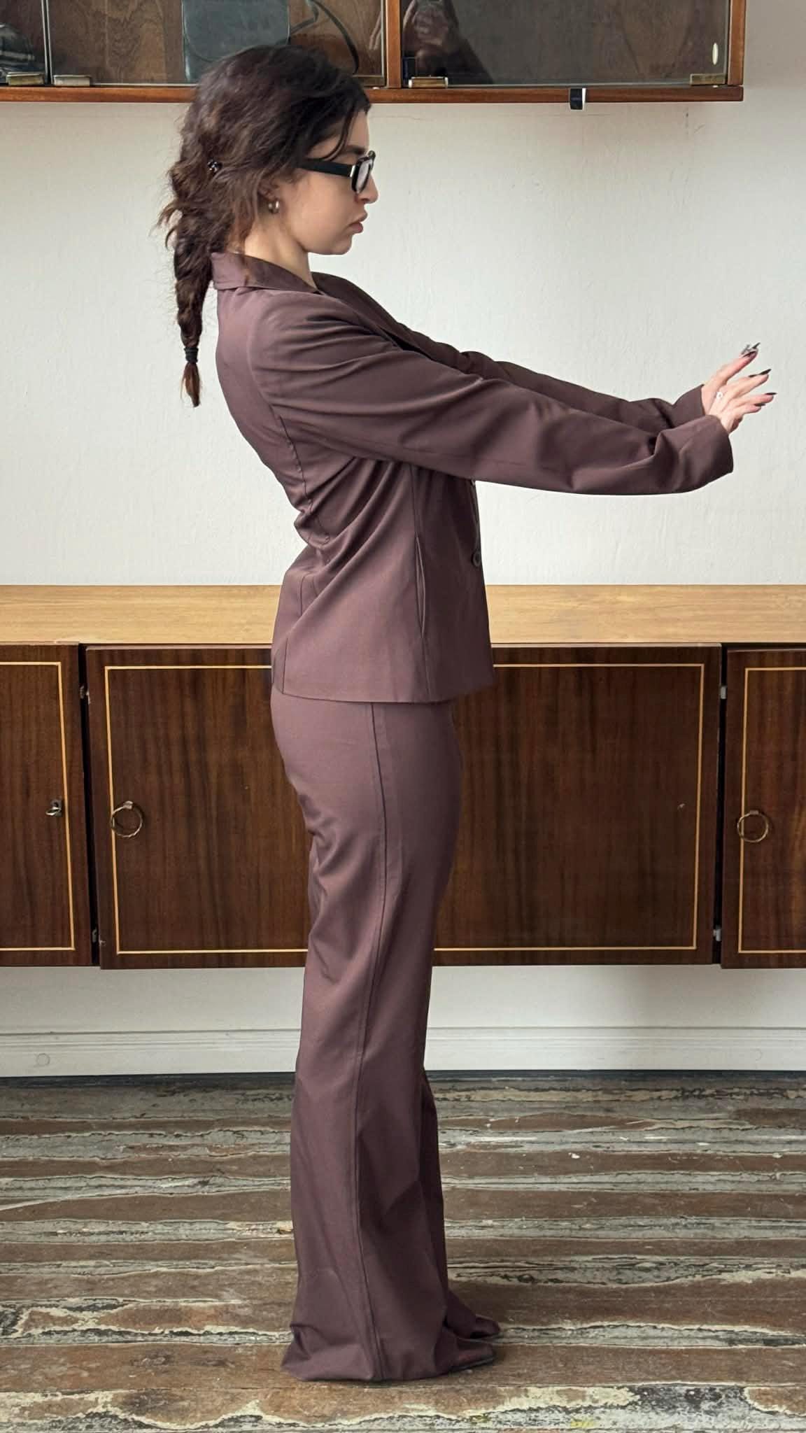 A woman in a brown suit stands with her arms outstretched, facing away from the camera.