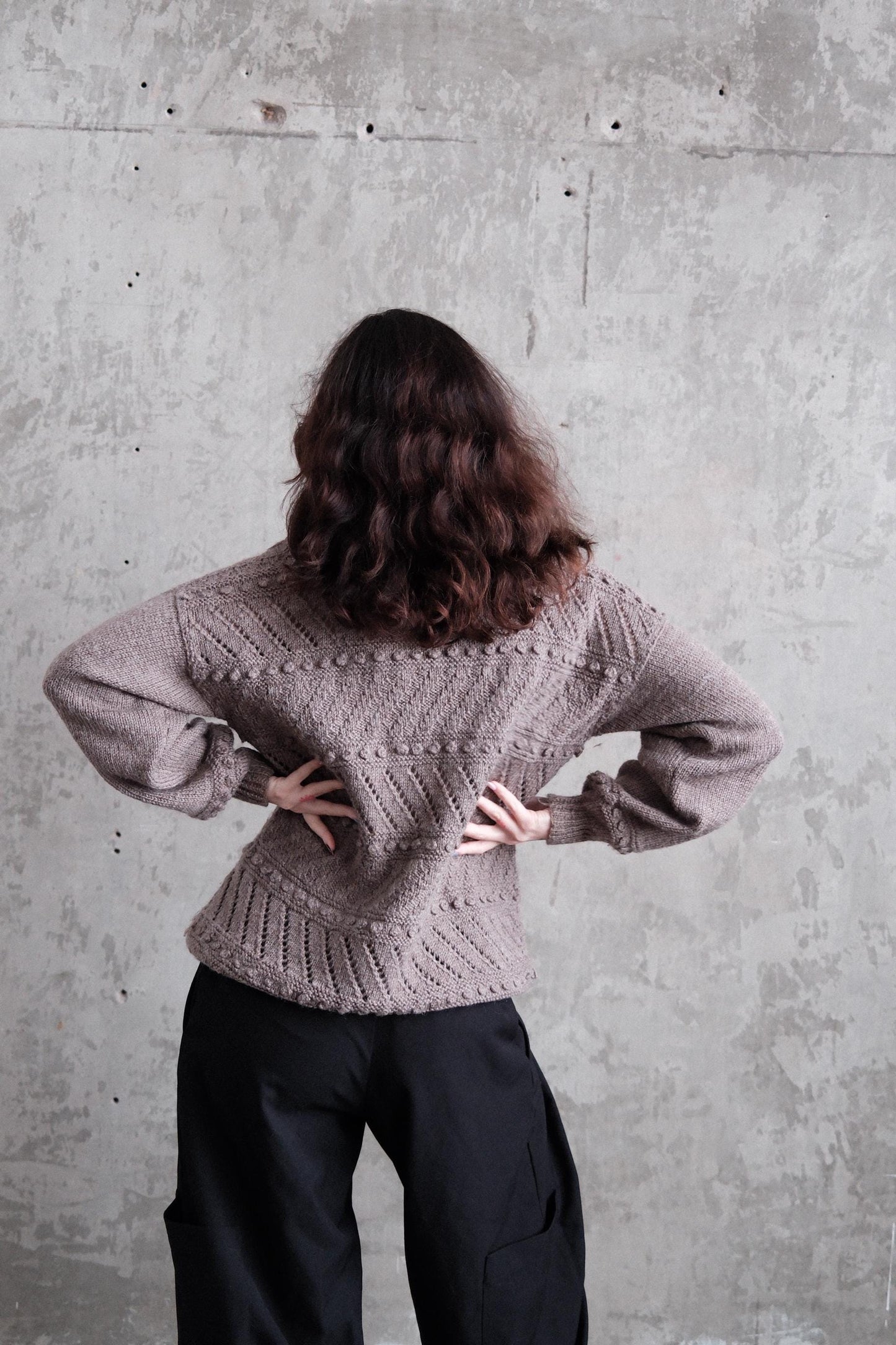 A person with long, curly hair is standing on a concrete floor, wearing a gray sweater and black pants.