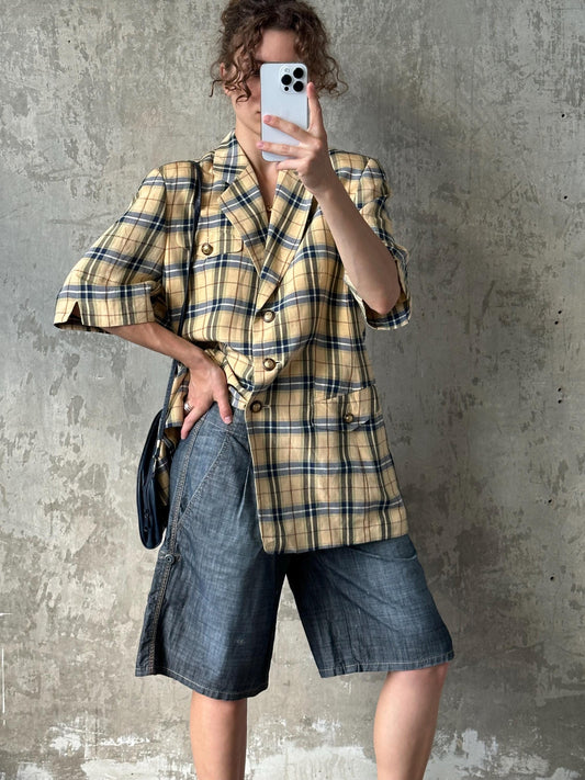 A person is taking a selfie with a smartphone, wearing a plaid jacket, denim shorts, and holding a bag.