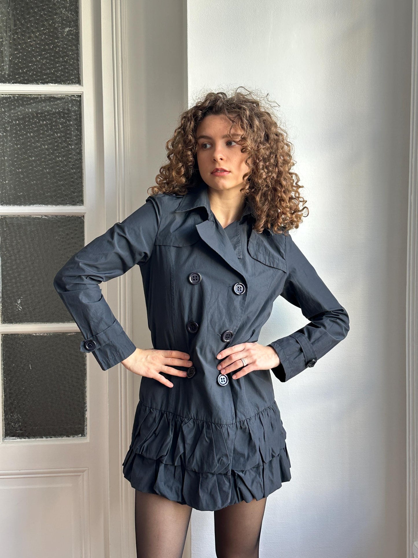 A woman with curly hair wearing a gray coat and skirt is posing in a relaxed manner.